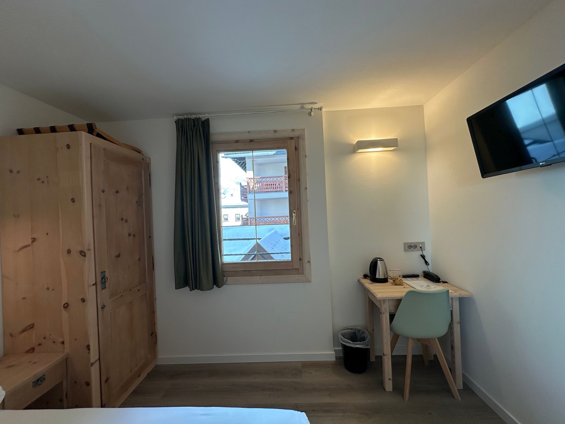 Camera da letto con armadio in legno, finestra con tenda, scrivania, sedia e TV a parete.