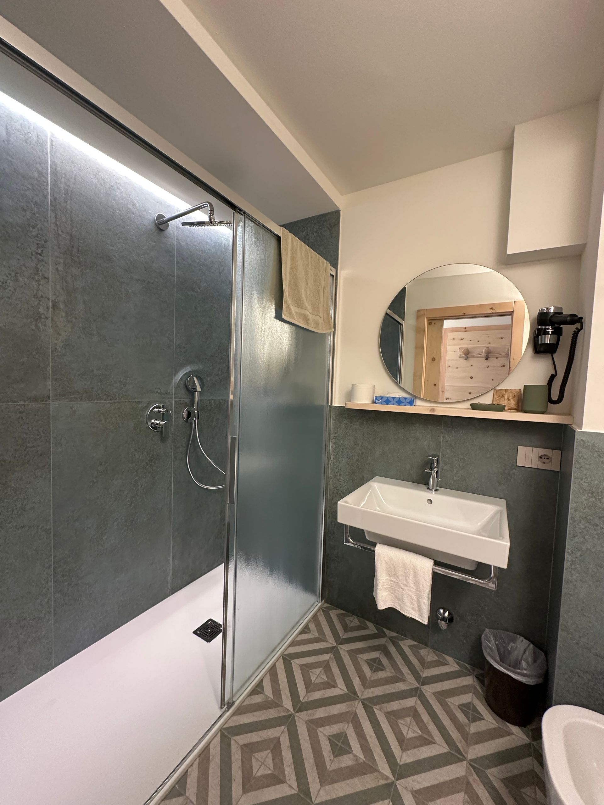 Bagno con doccia piastrellata grigia, lavandino, specchio rotondo e pavimento decorato.