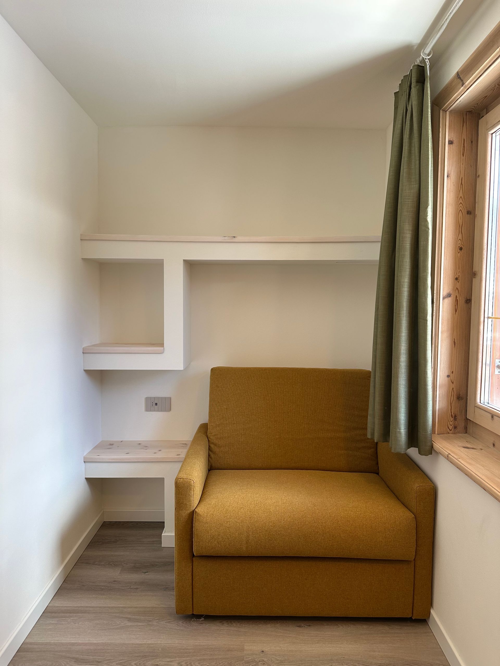 Piccola stanza con poltrona giallo senape, scaffali a muro, finestra con tenda verde. Pavimento in legno chiaro.