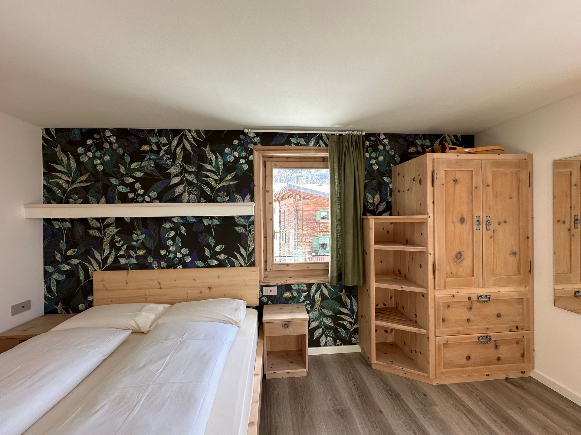 Camera da letto con carta da parati floreale, letto e armadio in legno, finestra con tenda verde.