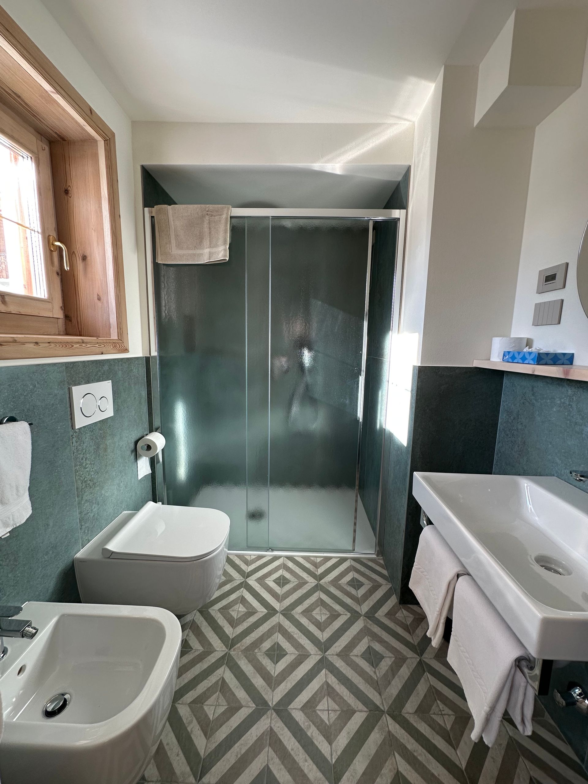 Bagno con WC, bidet, lavandino e doccia. Pareti e pavimento piastrellati in verde e grigio. Finestra sulla sinistra.