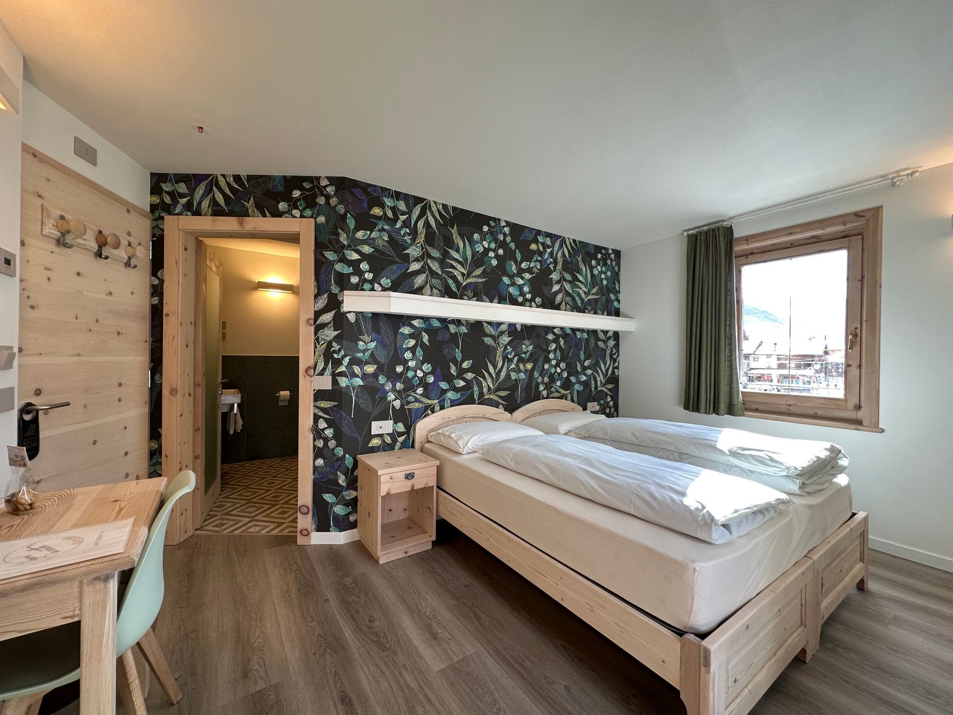 Camera da letto con mobili in legno, letto matrimoniale, carta da parati floreale, piccola finestra e bagno aperto.