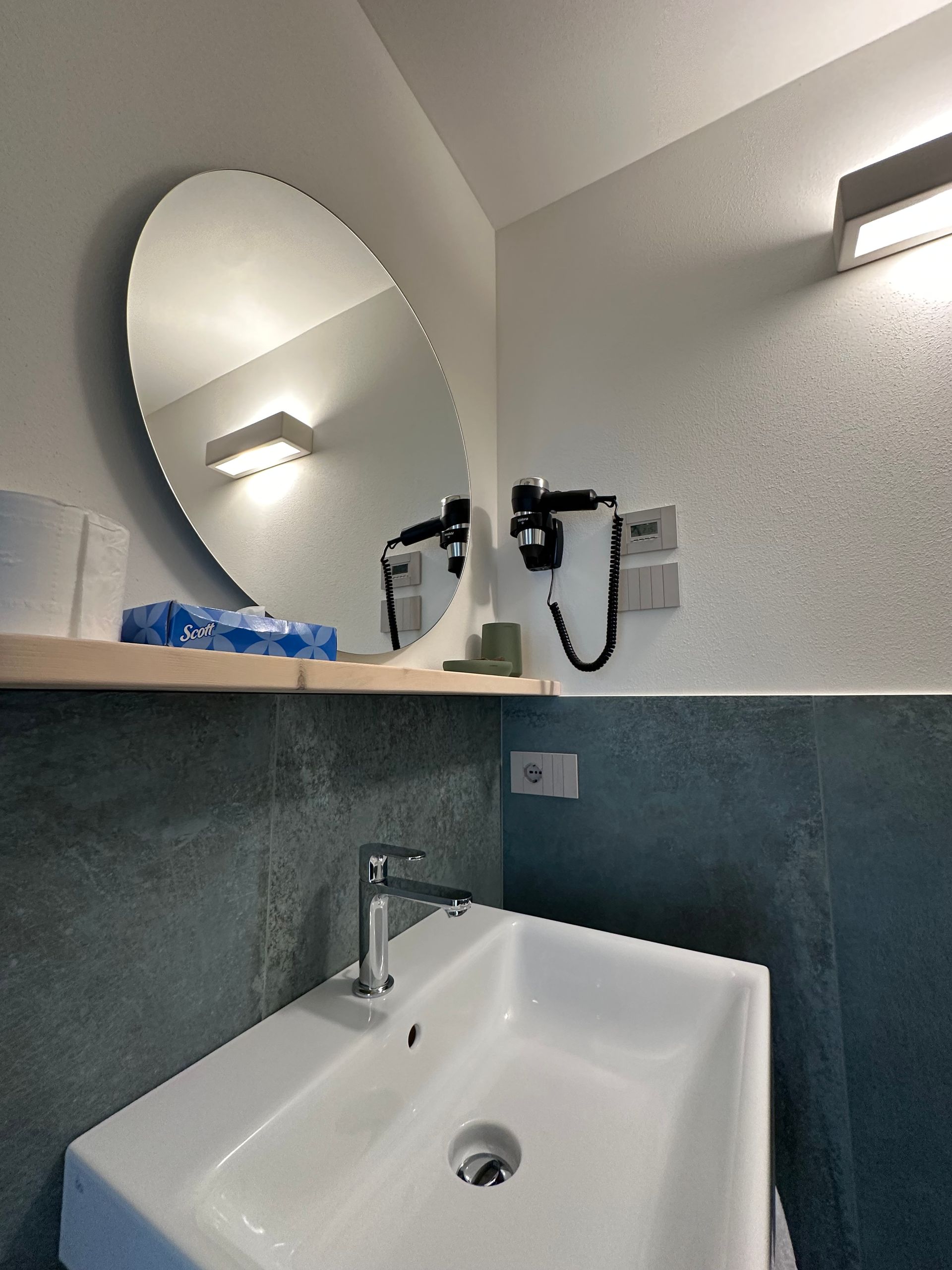Angolo del bagno con specchio ovale, lavandino bianco, asciugacapelli e pareti grigio-blu.