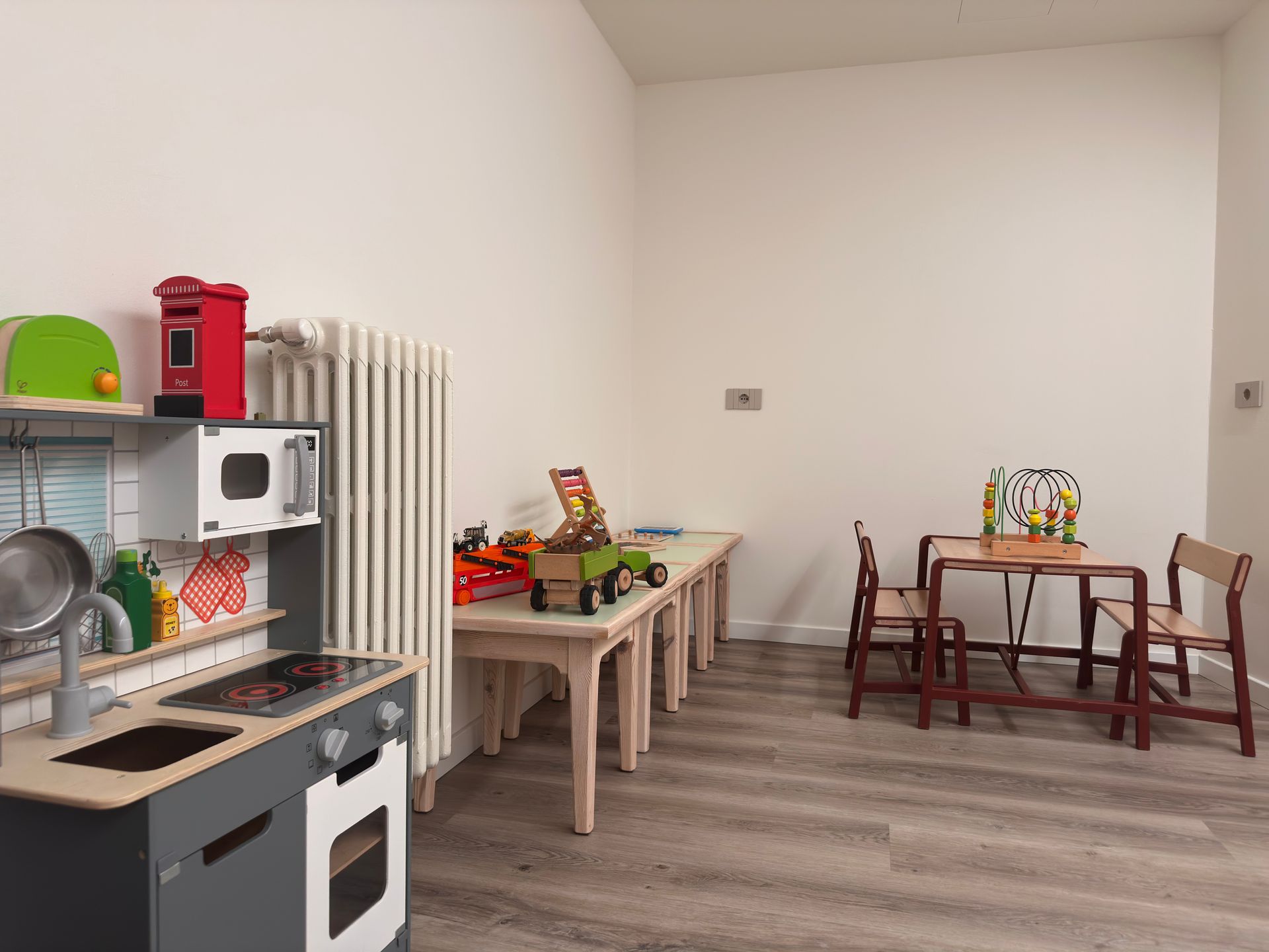 Un'area giochi con una cucina giocattolo, tavoli, sedie e vari giocattoli contro pareti bianche.