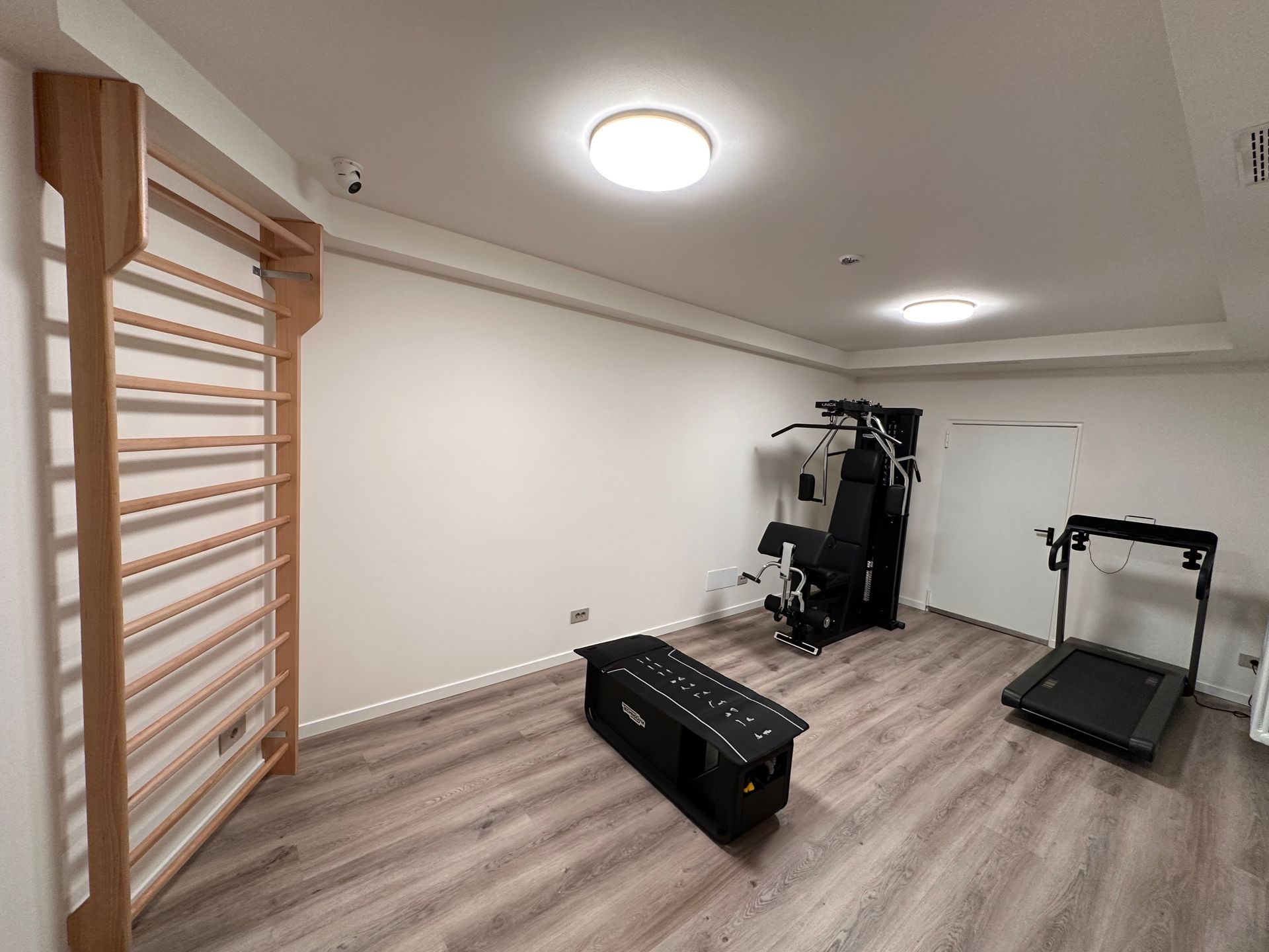 Interno della palestra con spalliera in legno, macchina per pesi, tapis roulant e panca nera su pavimento in legno chiaro.