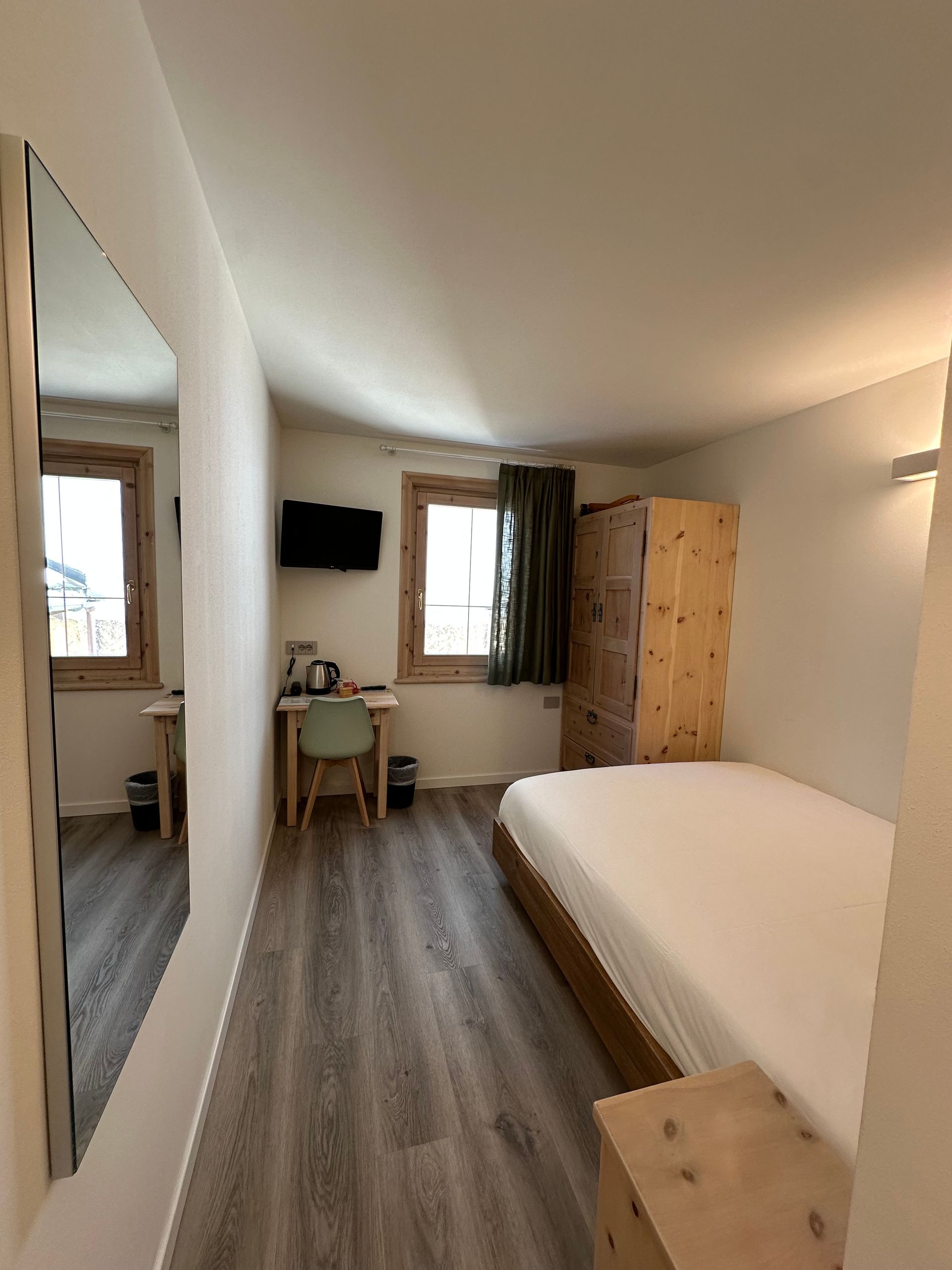 Camera da letto con letto, scrivania, armadio, finestra e specchio. Mobili e pavimenti in legno chiaro.