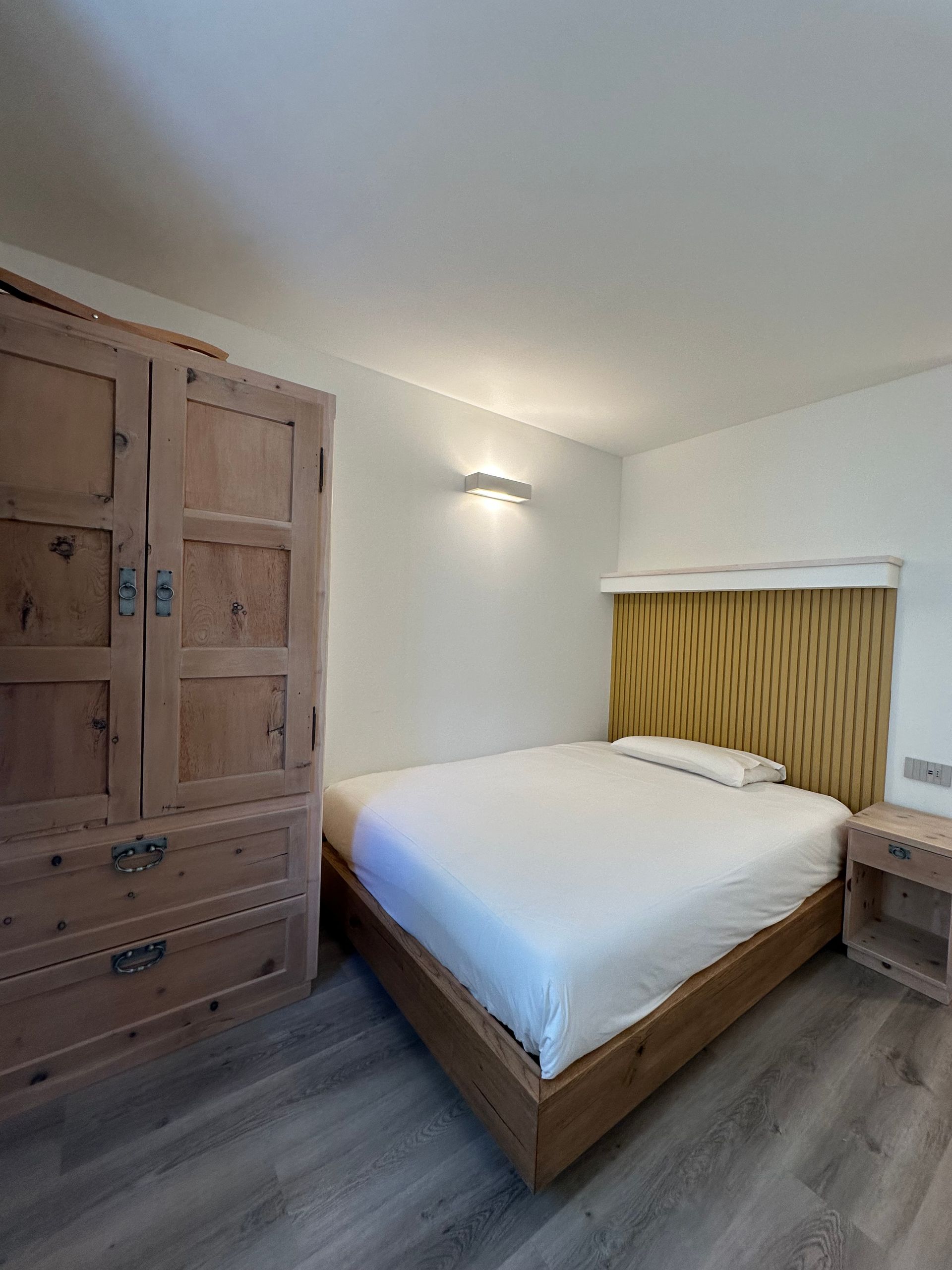 Camera da letto con mobili in legno: armadio, letto, comodino. Pareti bianche, pavimento grigio, toni neutri.