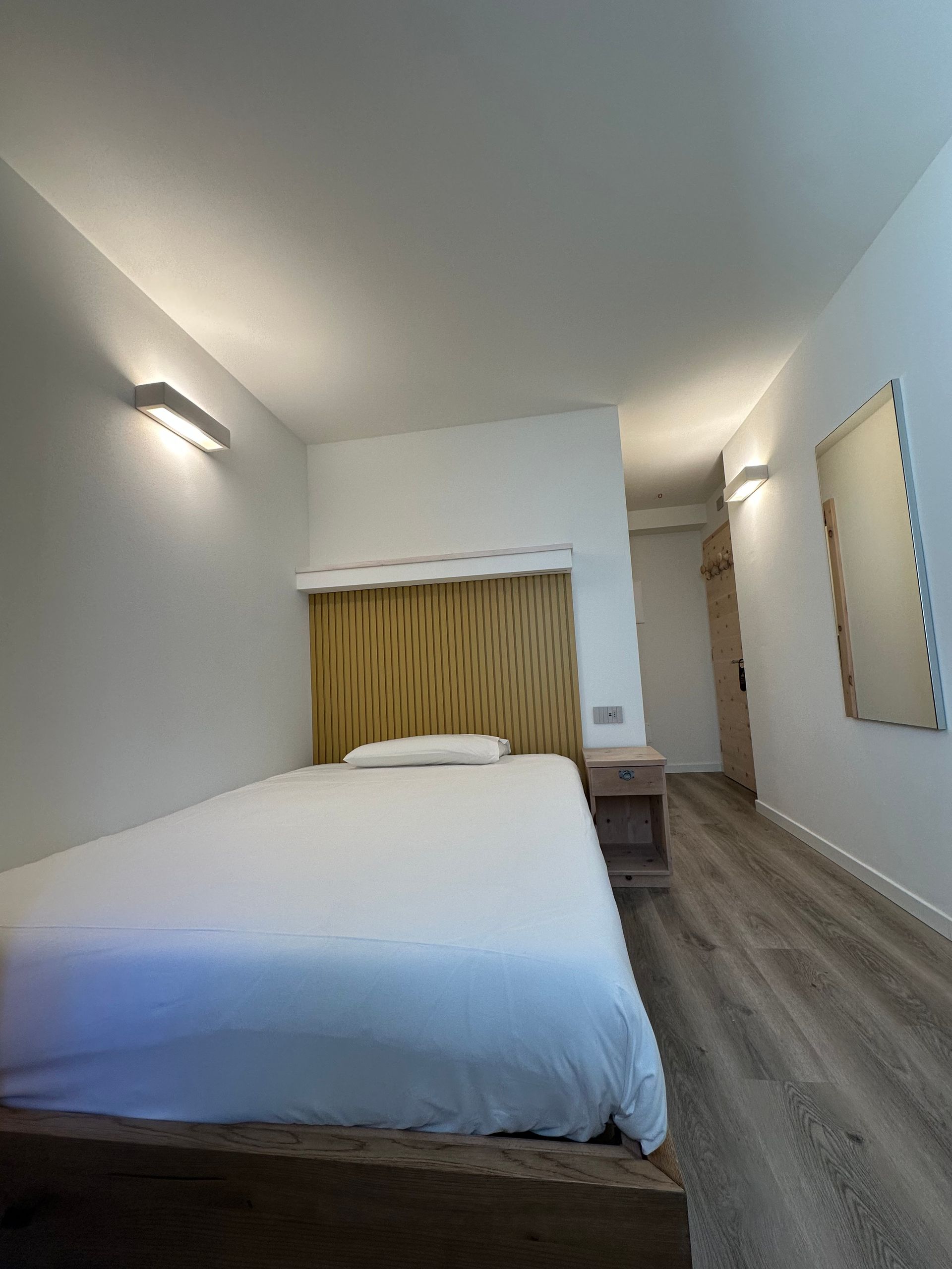 Camera da letto con letto, comodino, specchio e pavimento in legno. Le pareti sono bianche e c'è una parete decorativa con motivo in legno.