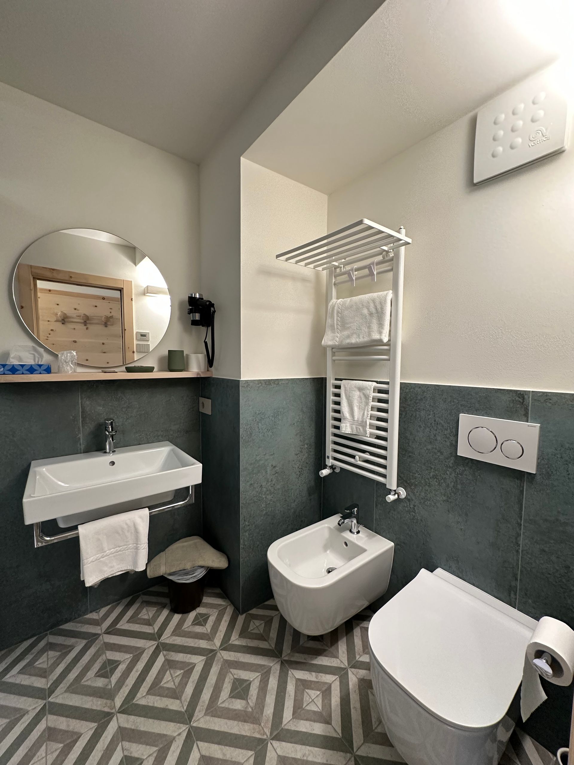 Bagno con pareti piastrellate color verde acqua, sanitari bianchi, specchio rotondo e pavimento decorato.