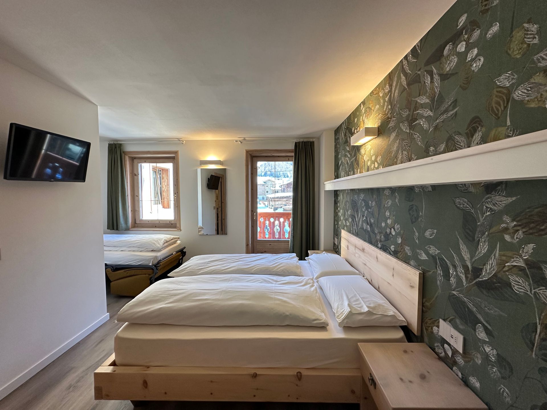 Camera da letto con un letto grande, carta da parati floreale, TV e un letto più piccolo sullo sfondo. Le finestre offrono una vista.