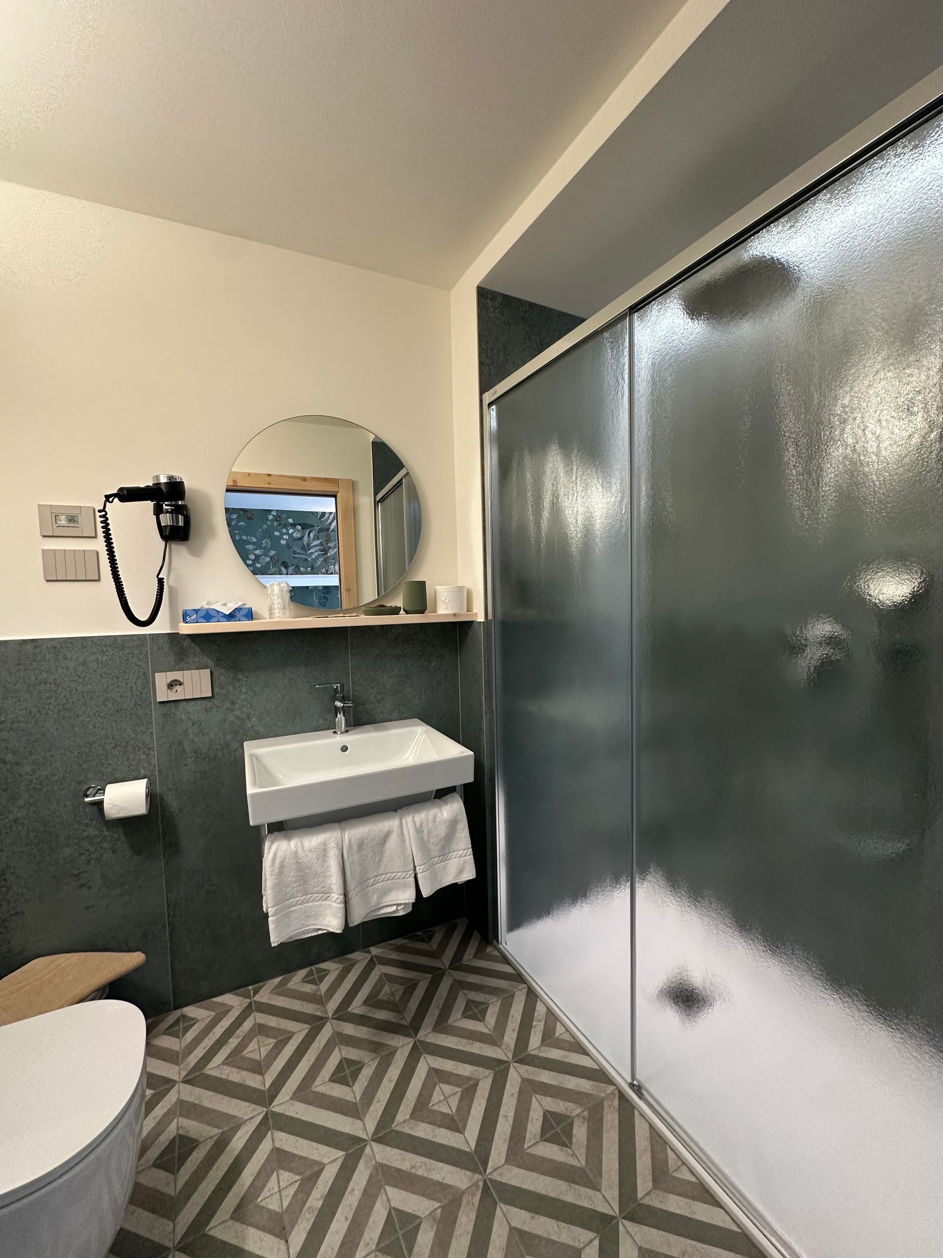 Bagno con doccia in vetro smerigliato, lavandino, WC e pavimento in piastrelle fantasia.