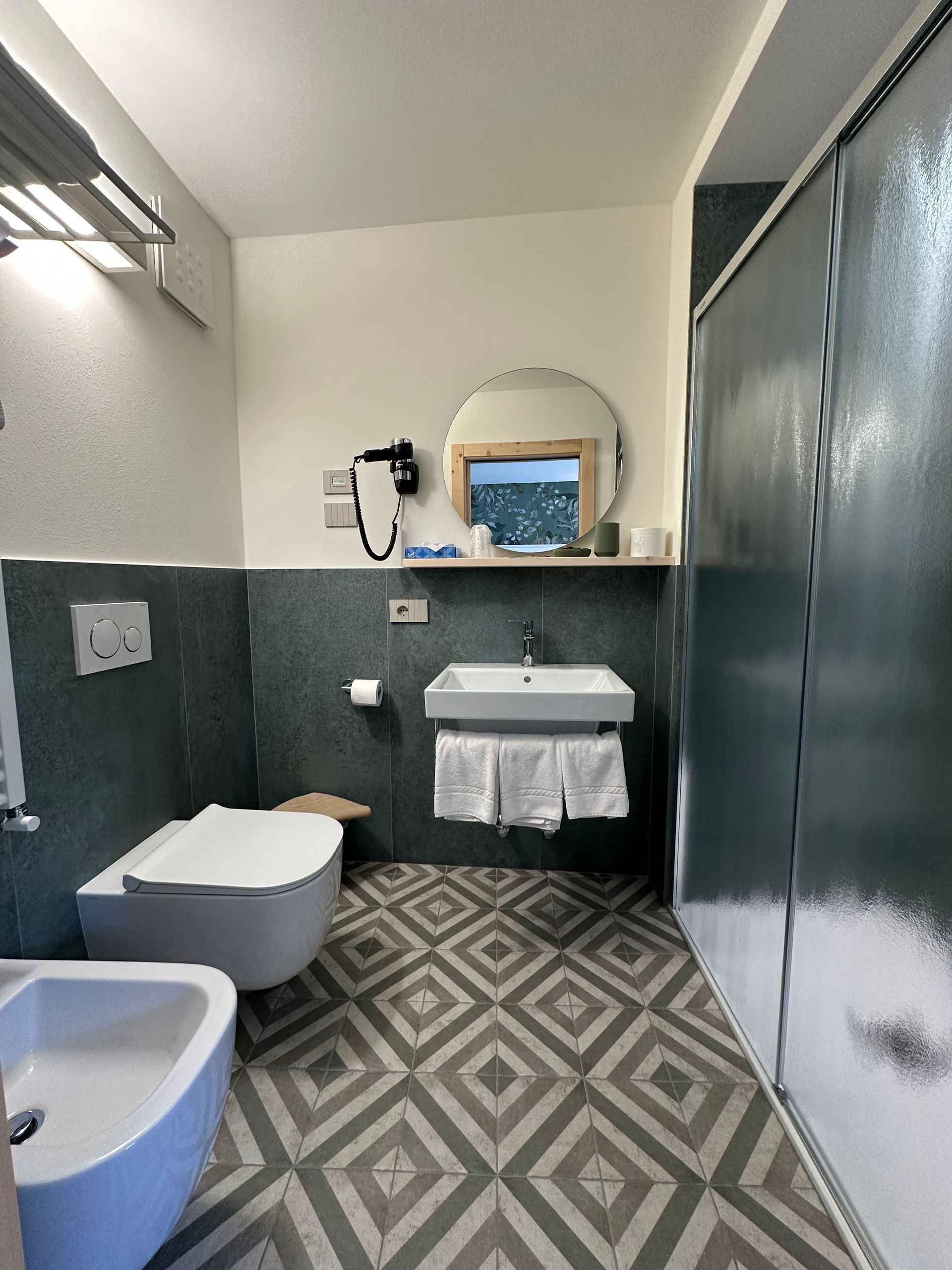 Bagno con WC, lavandino e doccia. Piastrelle blu e grigie, specchio circolare, portasciugamani.