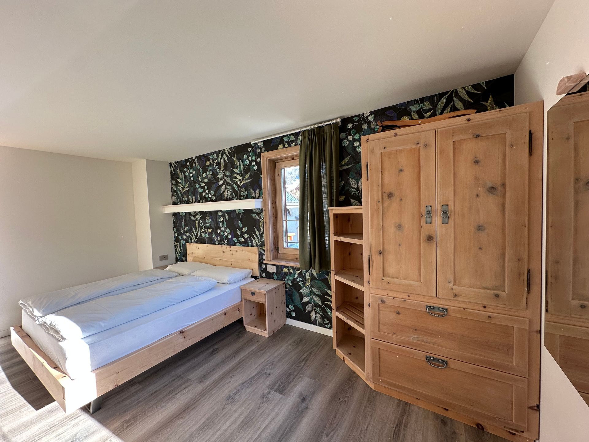 Camera da letto con mobili in legno chiaro, tra cui un letto, un comodino e un armadio contro una parete con carta da parati floreale.