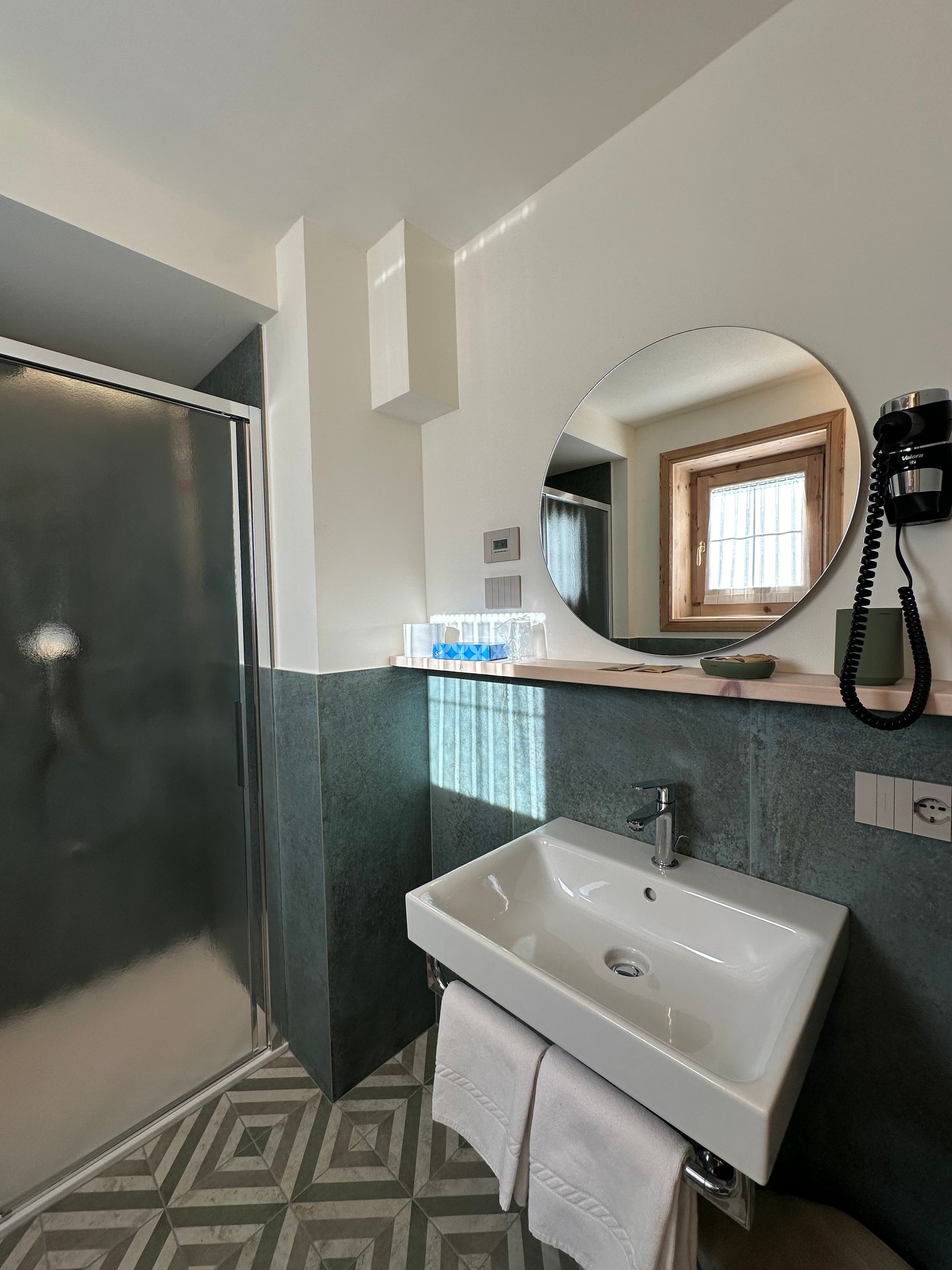 Bagno con doccia, lavandino e specchio rotondo. Pareti piastrellate color verde acqua, pavimento geometrico, riflesso nella finestra.