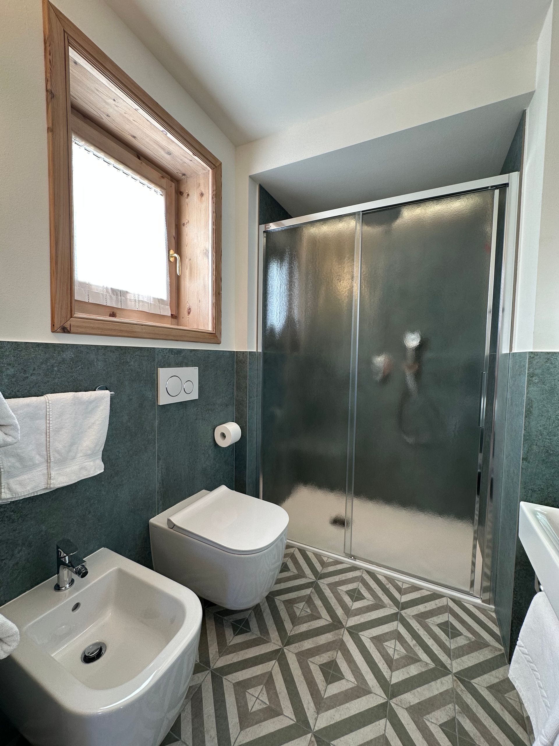 Bagno con WC, bidet, doccia, pavimento decorato, finestra e asciugamani a parete.