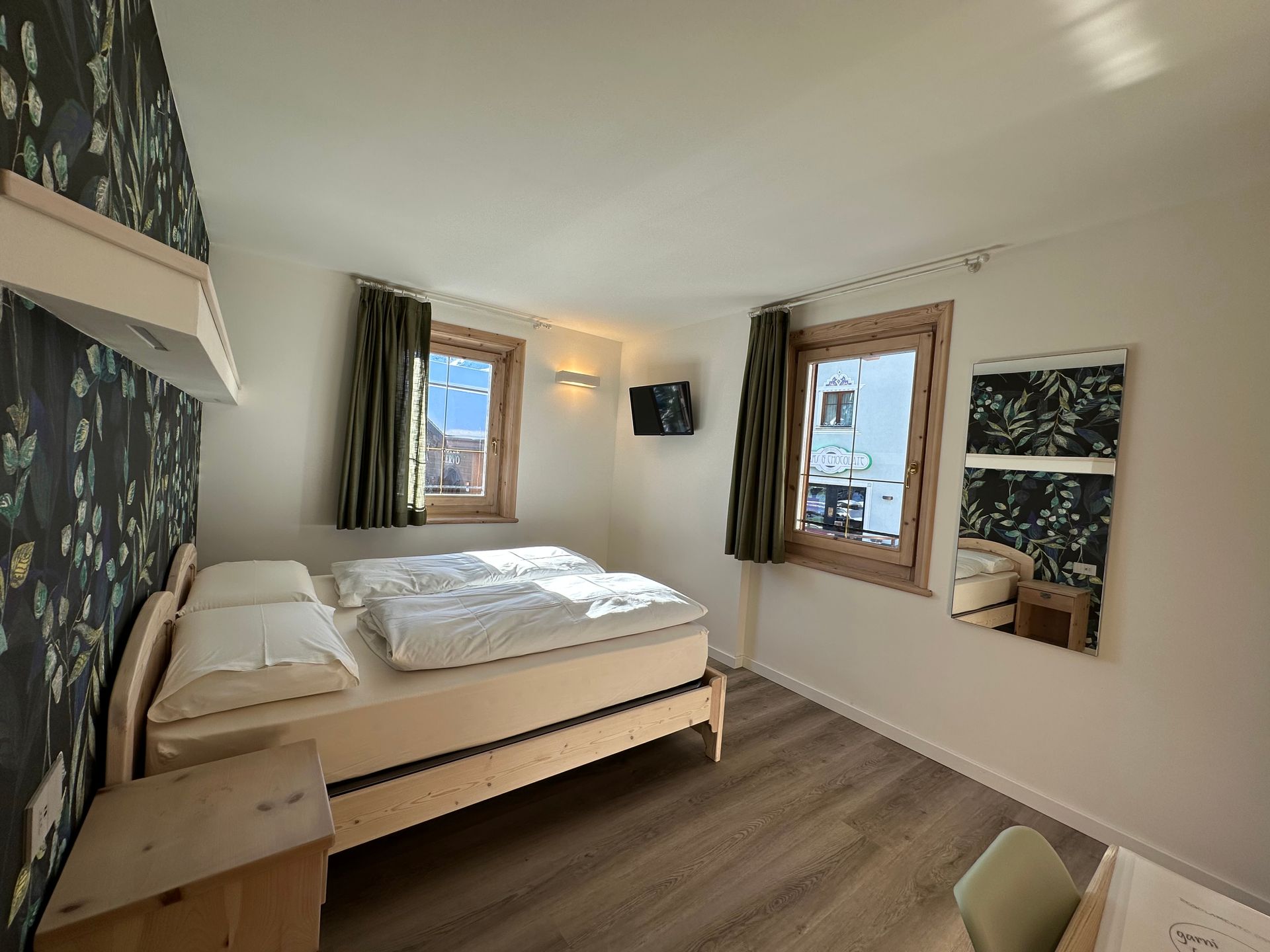 Camera da letto con letto in legno chiaro, due finestre, carta da parati a motivi e specchio, pavimento in legno.