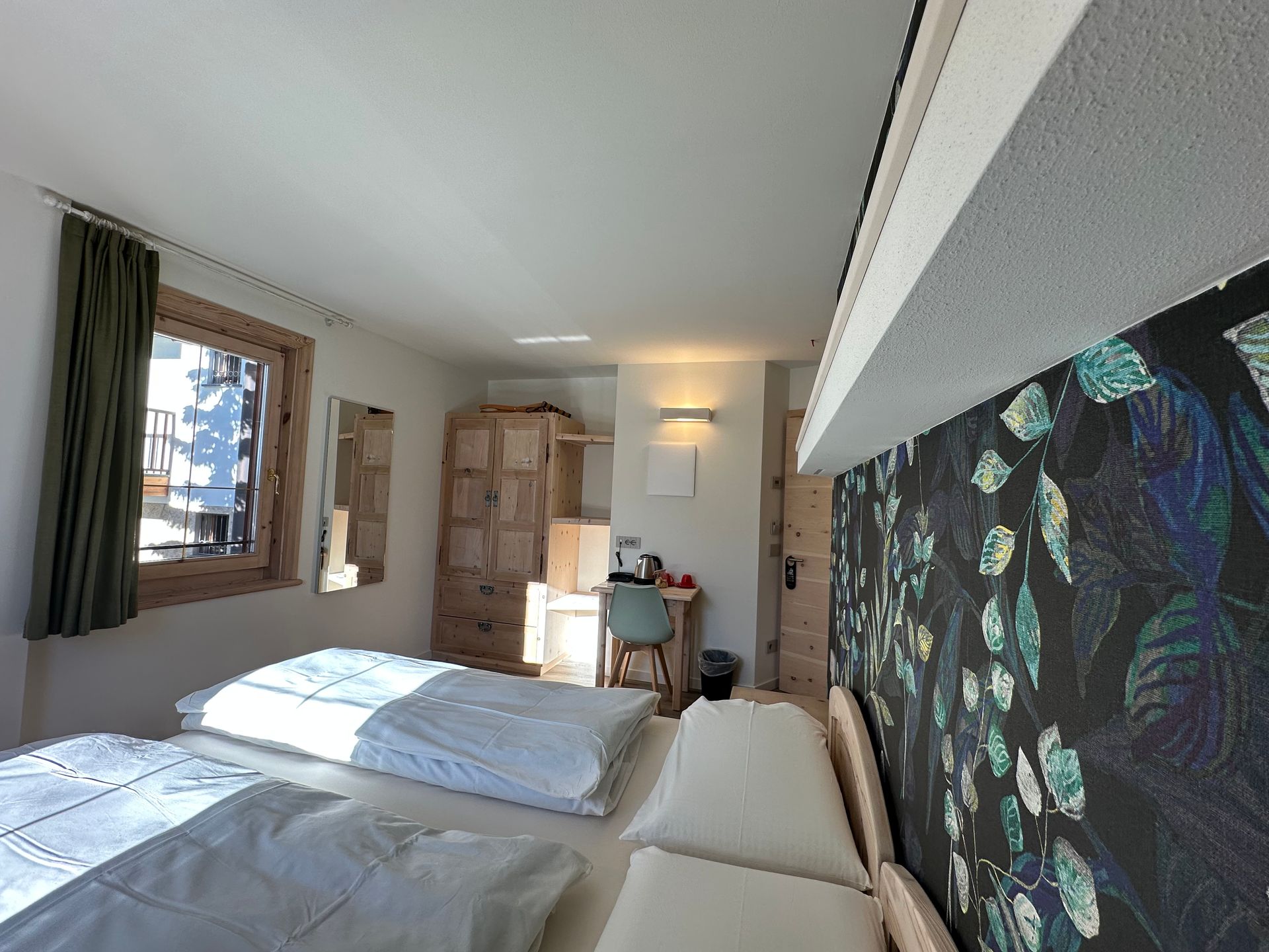 Camera da letto con due letti, mobili in legno e carta da parati a motivi. La luce del sole filtra attraverso una finestra.