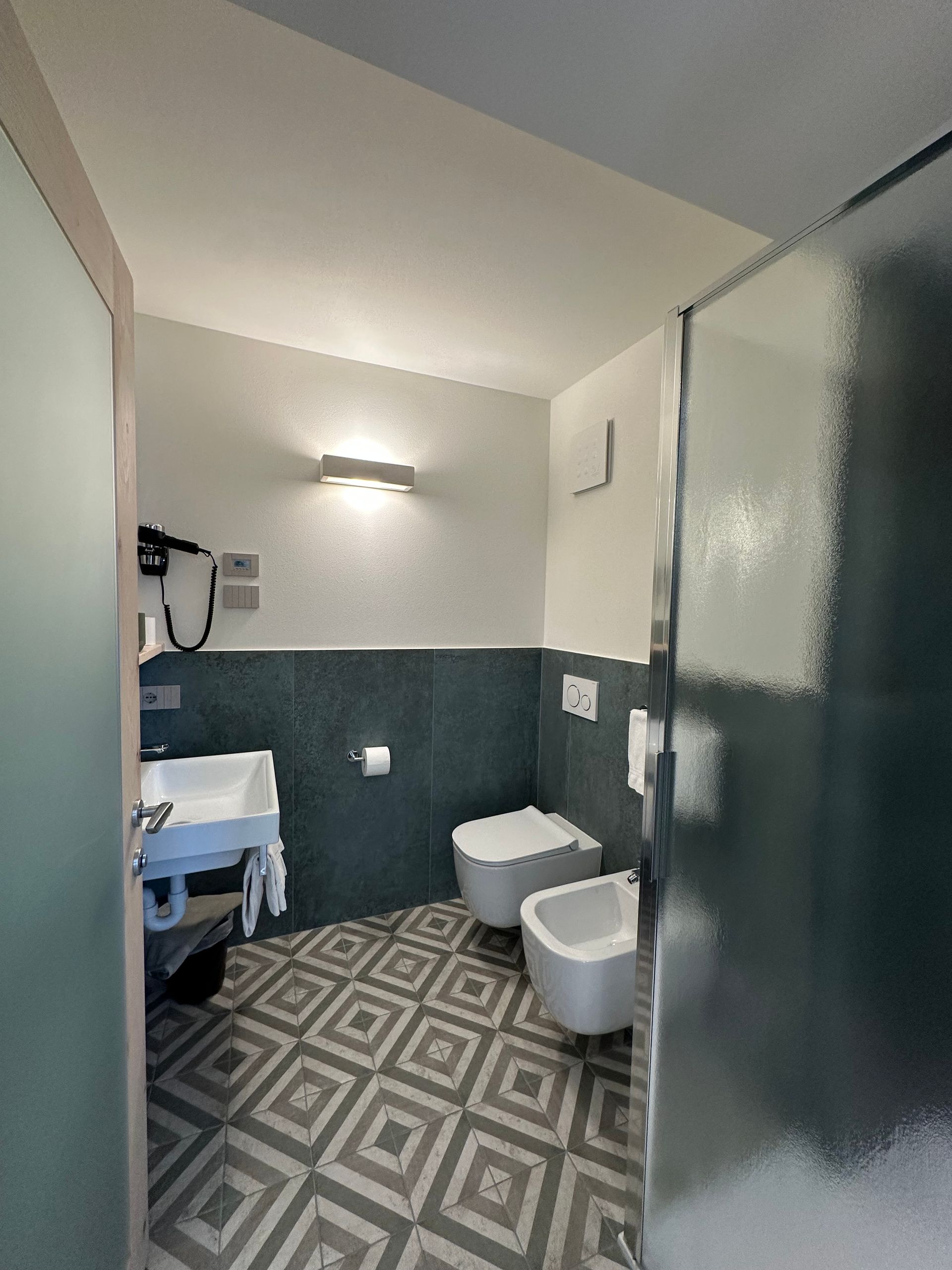 Bagno con pavimento decorato, piastrelle blu-verdi, WC, bidet, lavandino e porta della doccia in vetro smerigliato.