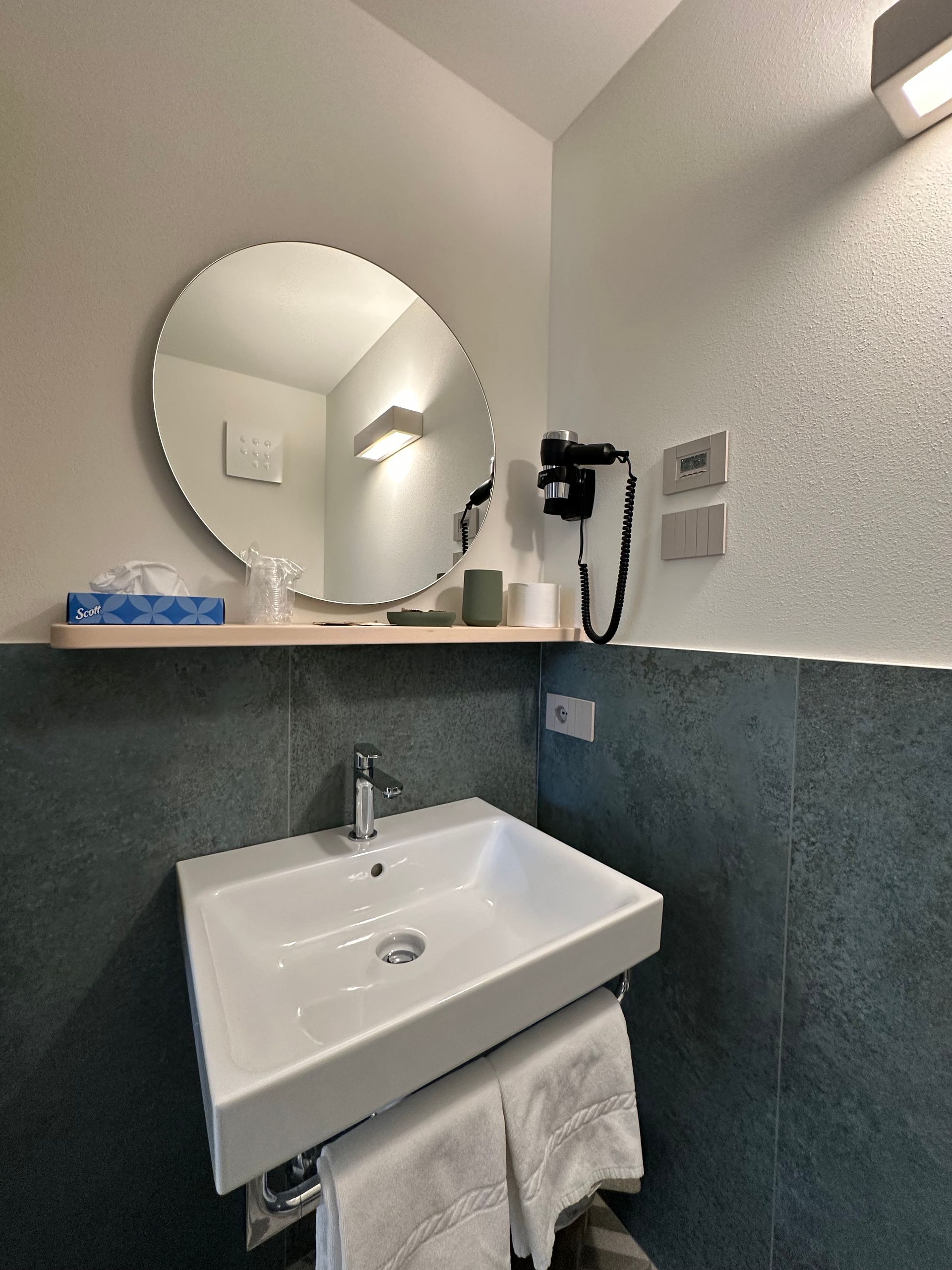 Bagno con lavandino bianco, specchio rotondo e piastrelle color verde acqua.