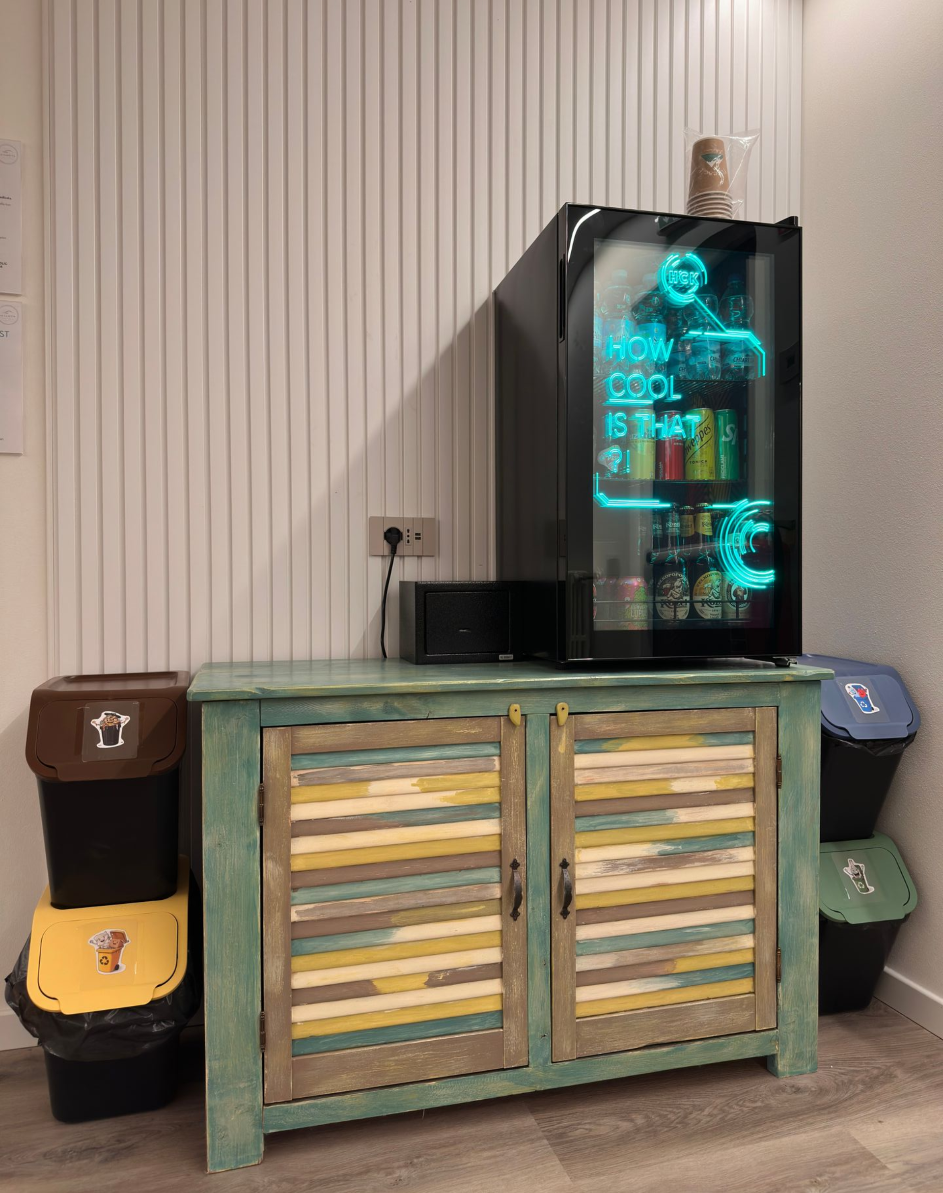 Minifrigo con luce blu brillante su un mobiletto verde acqua, accanto a colorati cestini della spazzatura.