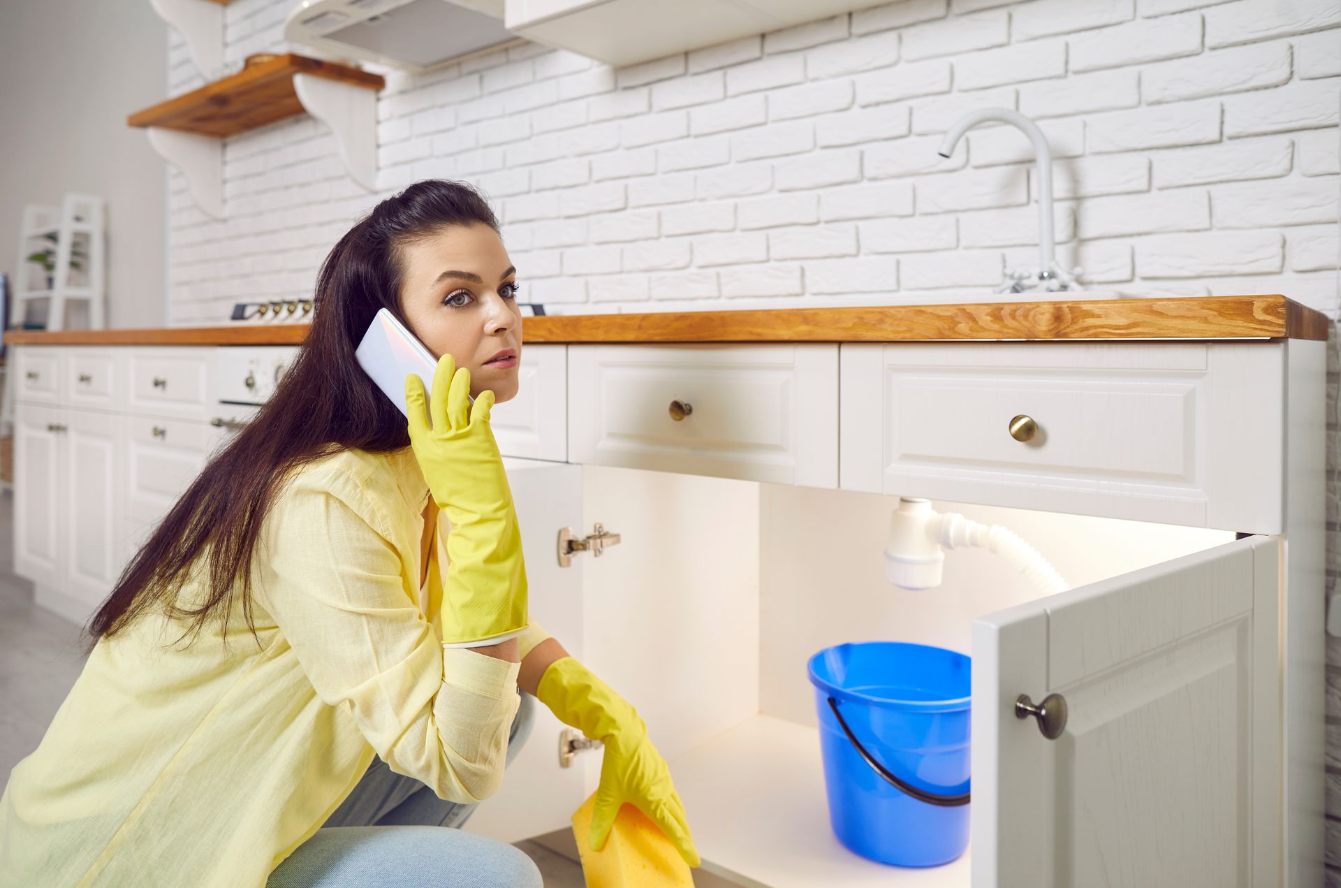 A woman crouching in front of an open kitchen cabinet with a blue bucket inside.