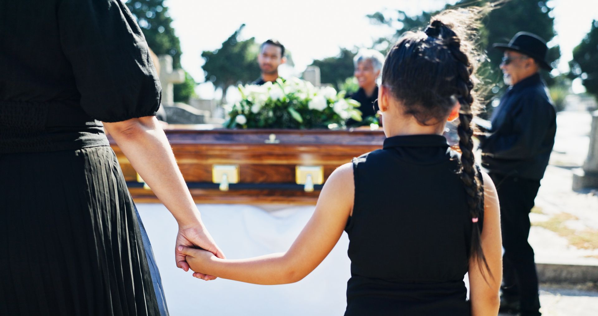 A woman and child in black at a funeral, holding hands near a coffin. Other mourners stand behind.