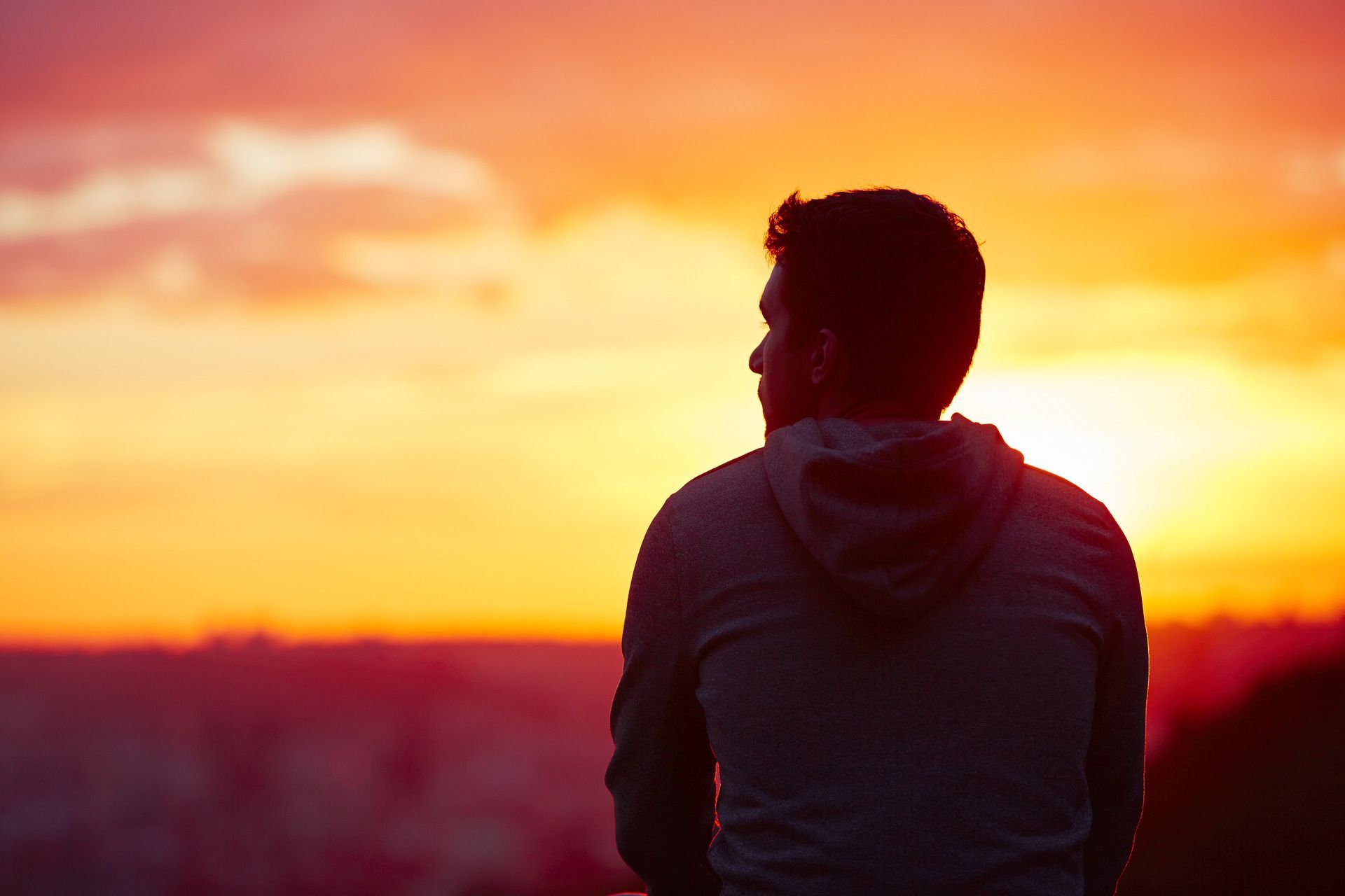 Man in hoodie silhouetted against a vibrant orange and pink sunset, looking out.