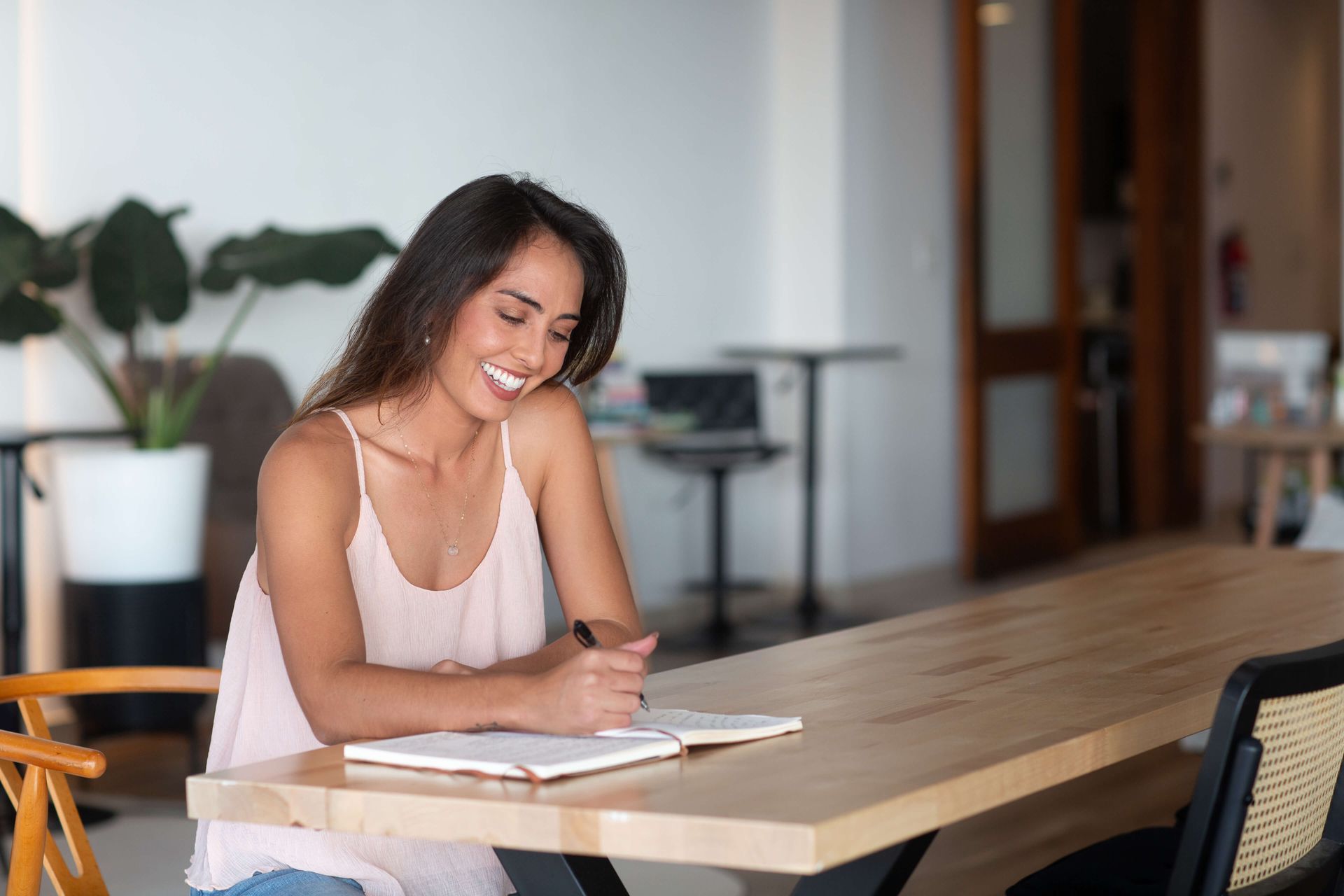 woman sitting at a coworking space wearing a pink tank top, smiling, and writing in a notebook.