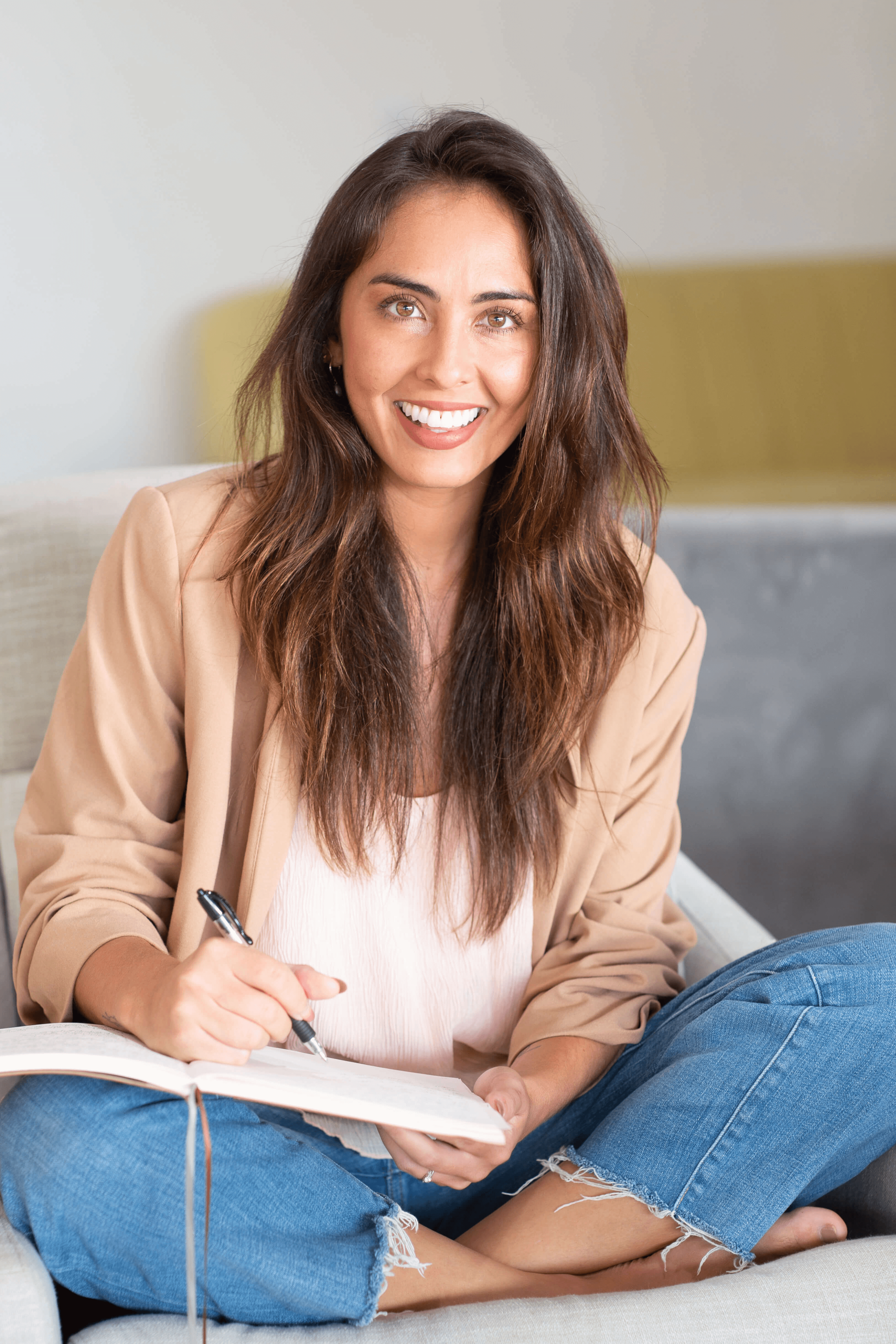 young woman smiling with long brown hair writing in a journal sitting in a large brown leather chair with white windows behind her