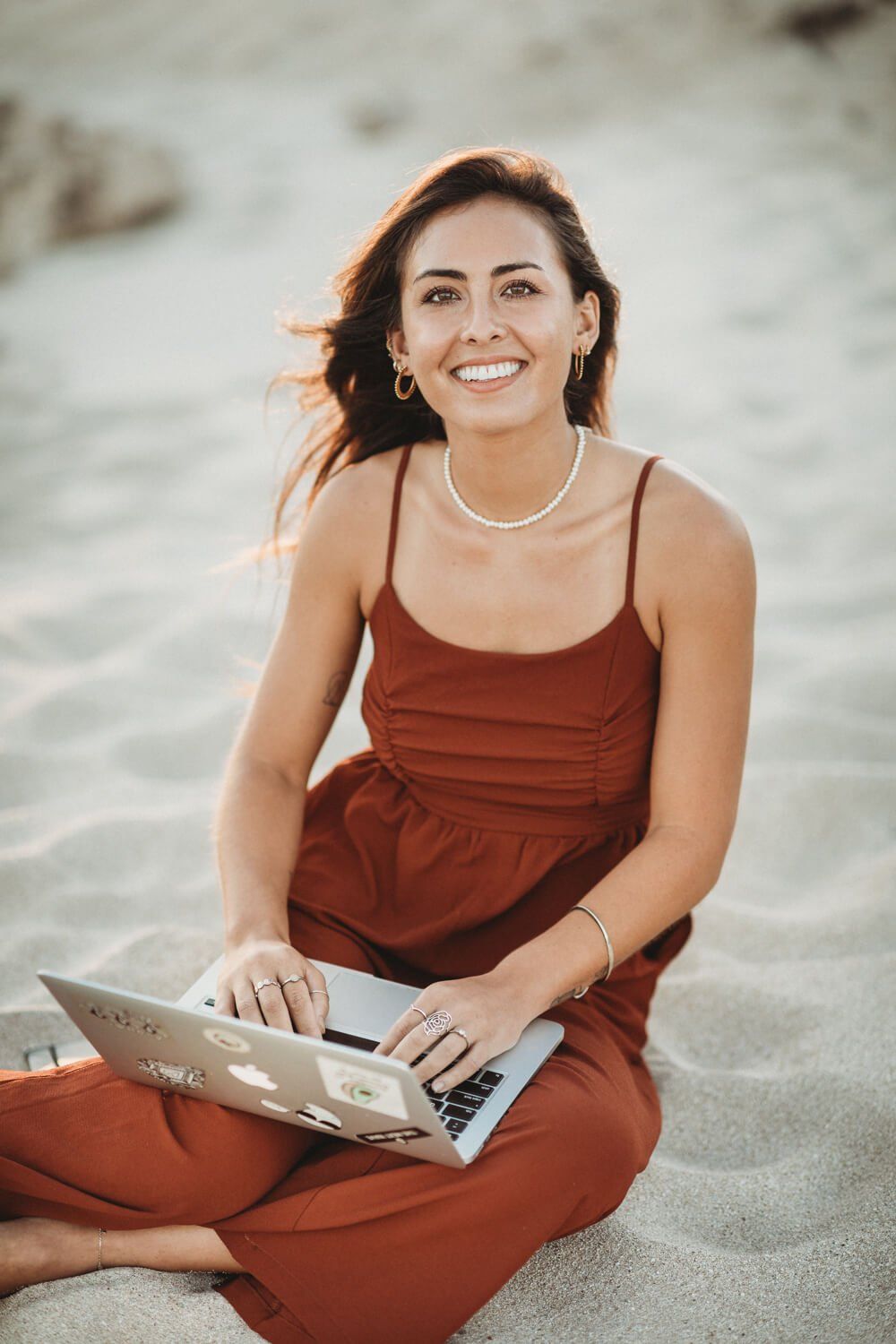 young woman sitting on the sand in a rust romper typing on a computer