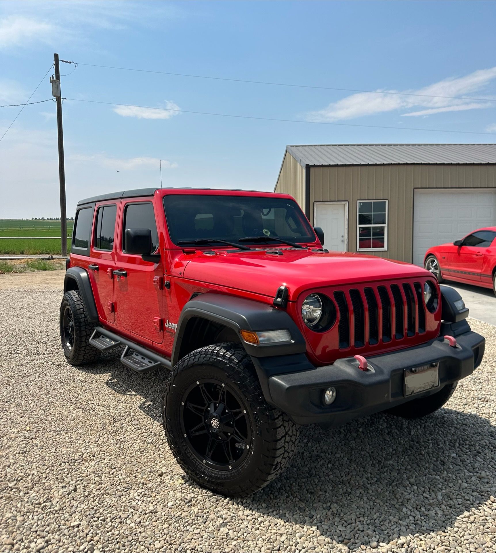 A red jeep is parked in a gravel lot in front of a garage.