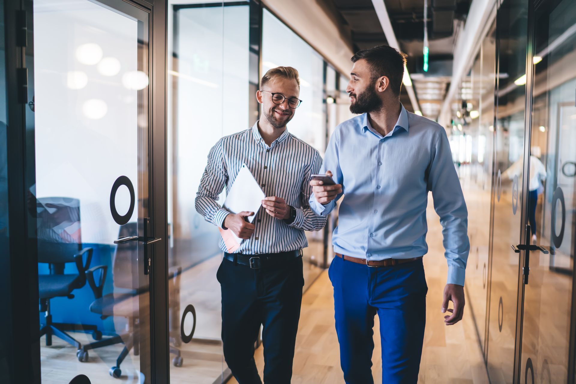 Two men are walking down a hallway in an office talking to each other.