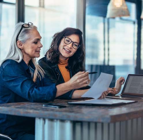 Two women are sitting at a table looking at papers and a laptop.