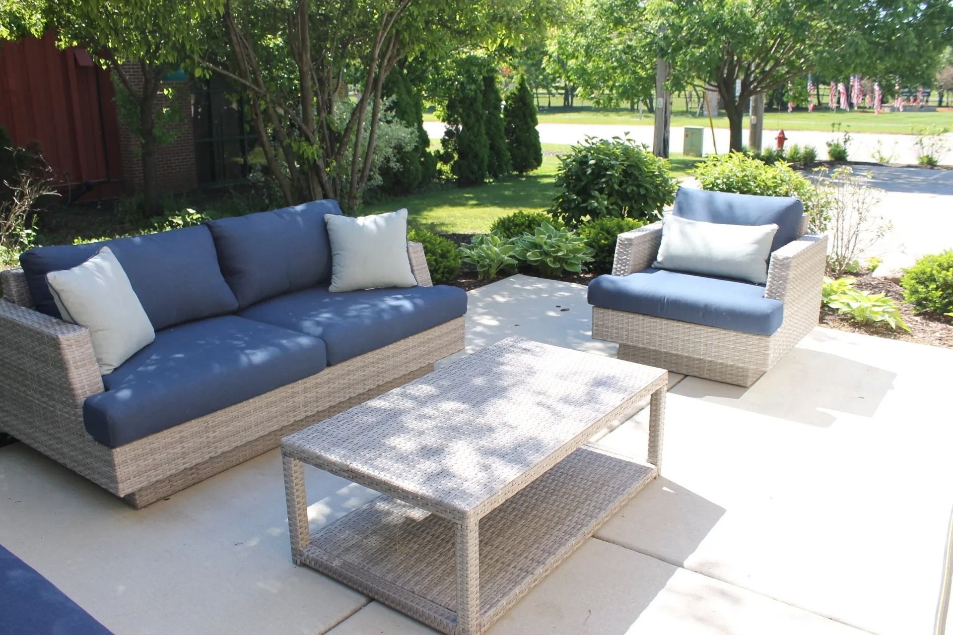 Outdoor seating area with blue cushions, gray wicker furniture, coffee table, and greenery.
