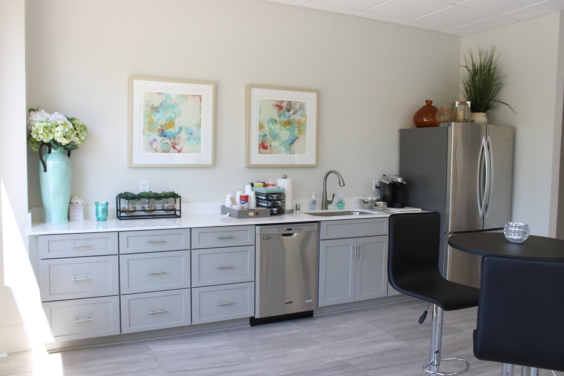 A small kitchenette with gray cabinets, dishwasher, sink, and fridge. Two paintings hang above a white countertop.