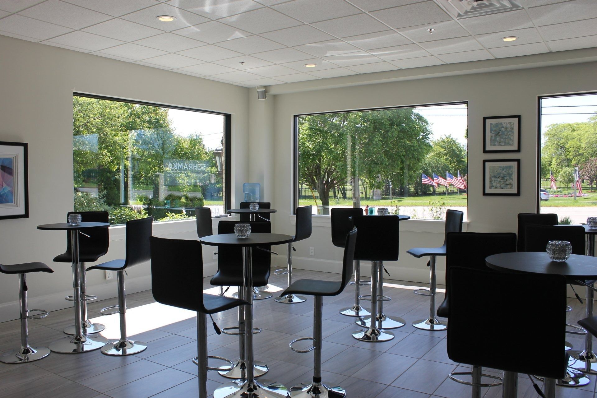 A room with tall, black chairs at small, round tables near large windows with an outdoor view.