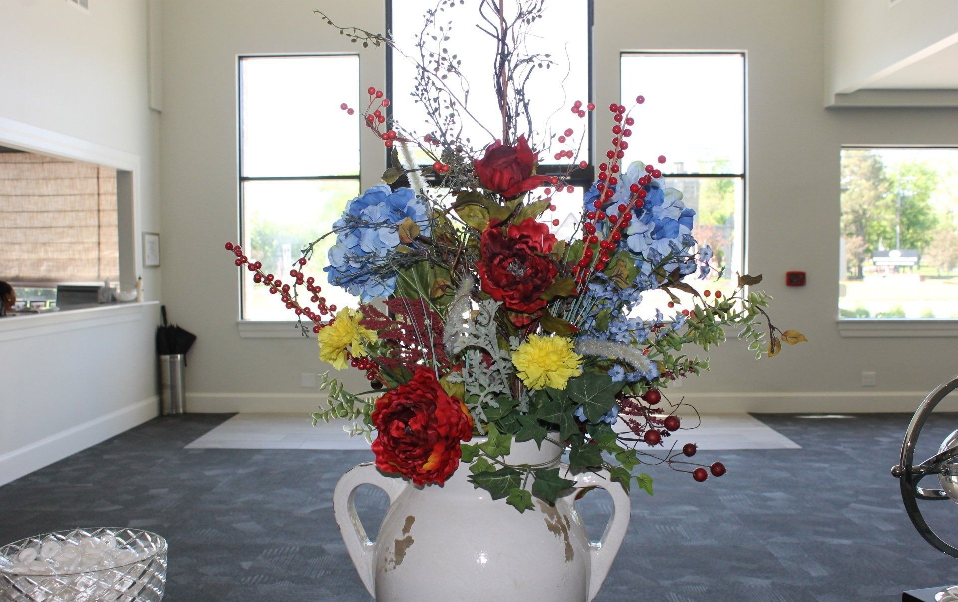 Large floral arrangement in white vase, in front of windows. Red, blue, and yellow flowers.