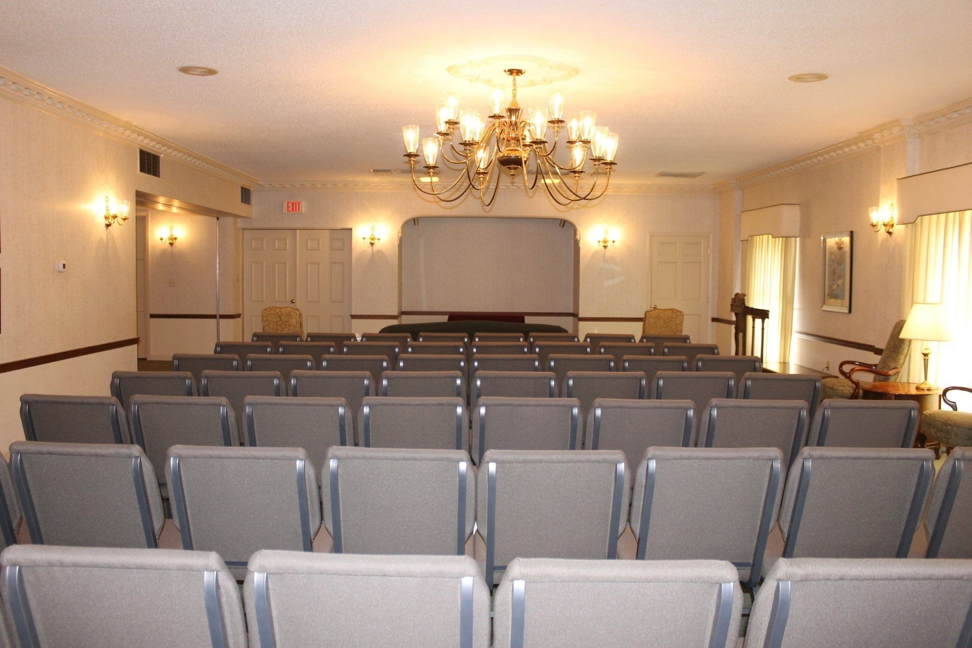Rows of gray chairs face a raised platform in a brightly lit room with a chandelier.
