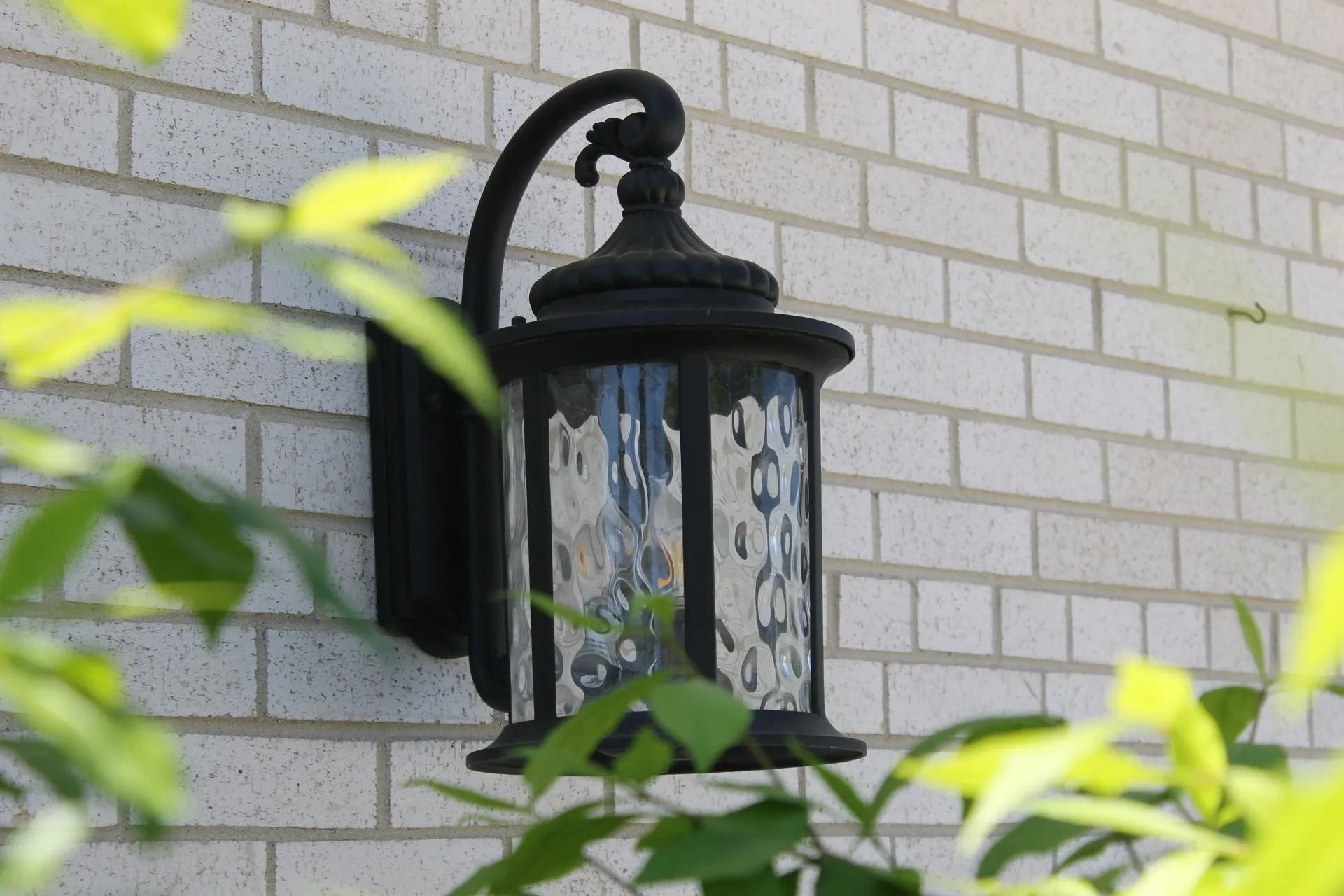 Black outdoor lantern on a brick wall, partially obscured by green leaves.