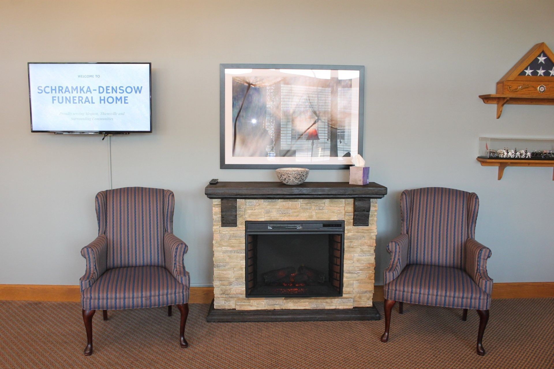 Lounge area at a funeral home with fireplace, chairs, and memorial displays on a gray wall.
