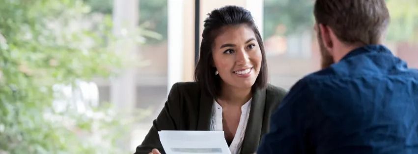 Woman in a blazer smiles and looks at a person as they sit at a table. A document is in front of them.