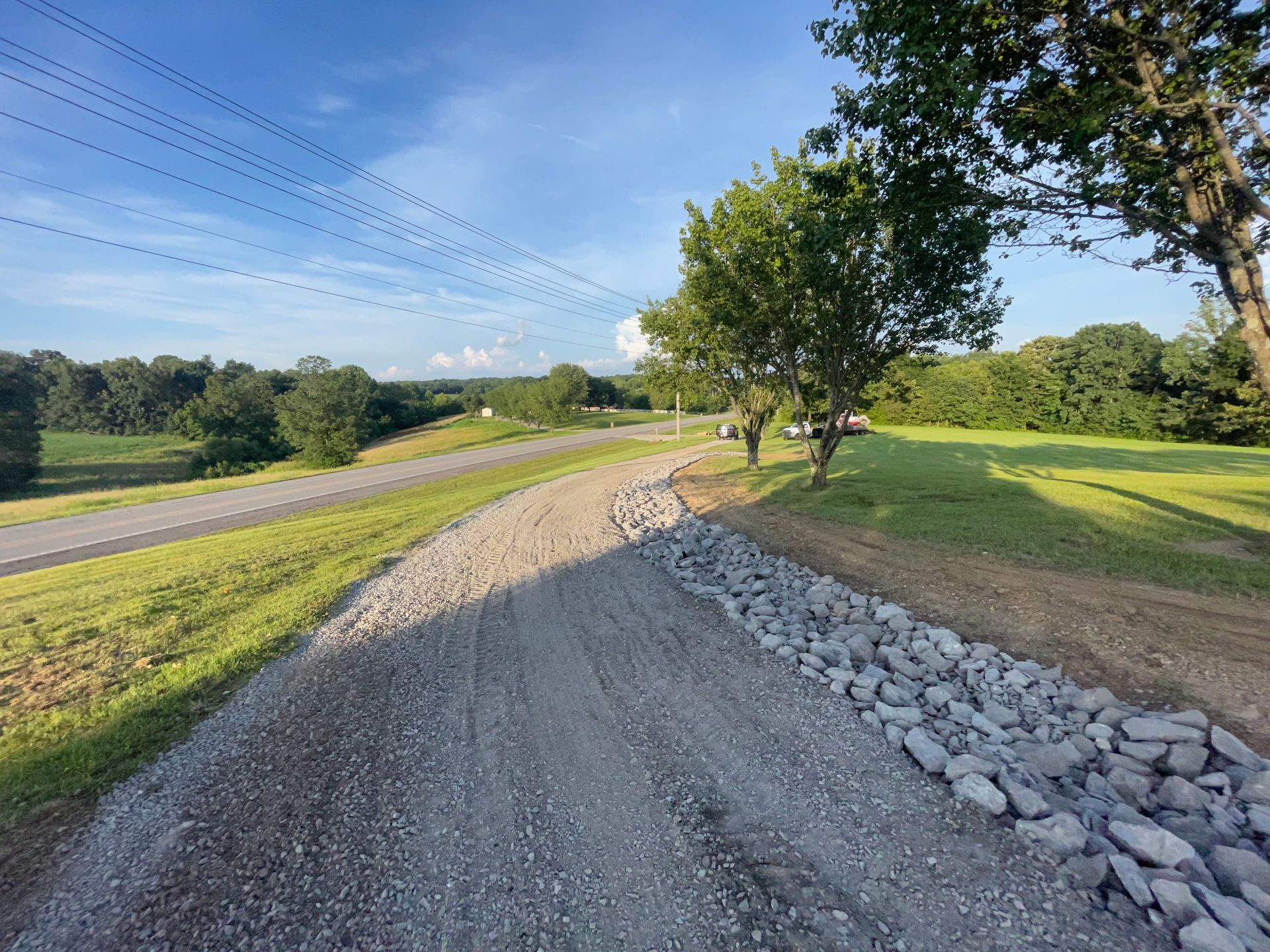 Gravel road alongside a grassy field, trees, and a paved road under a blue sky.