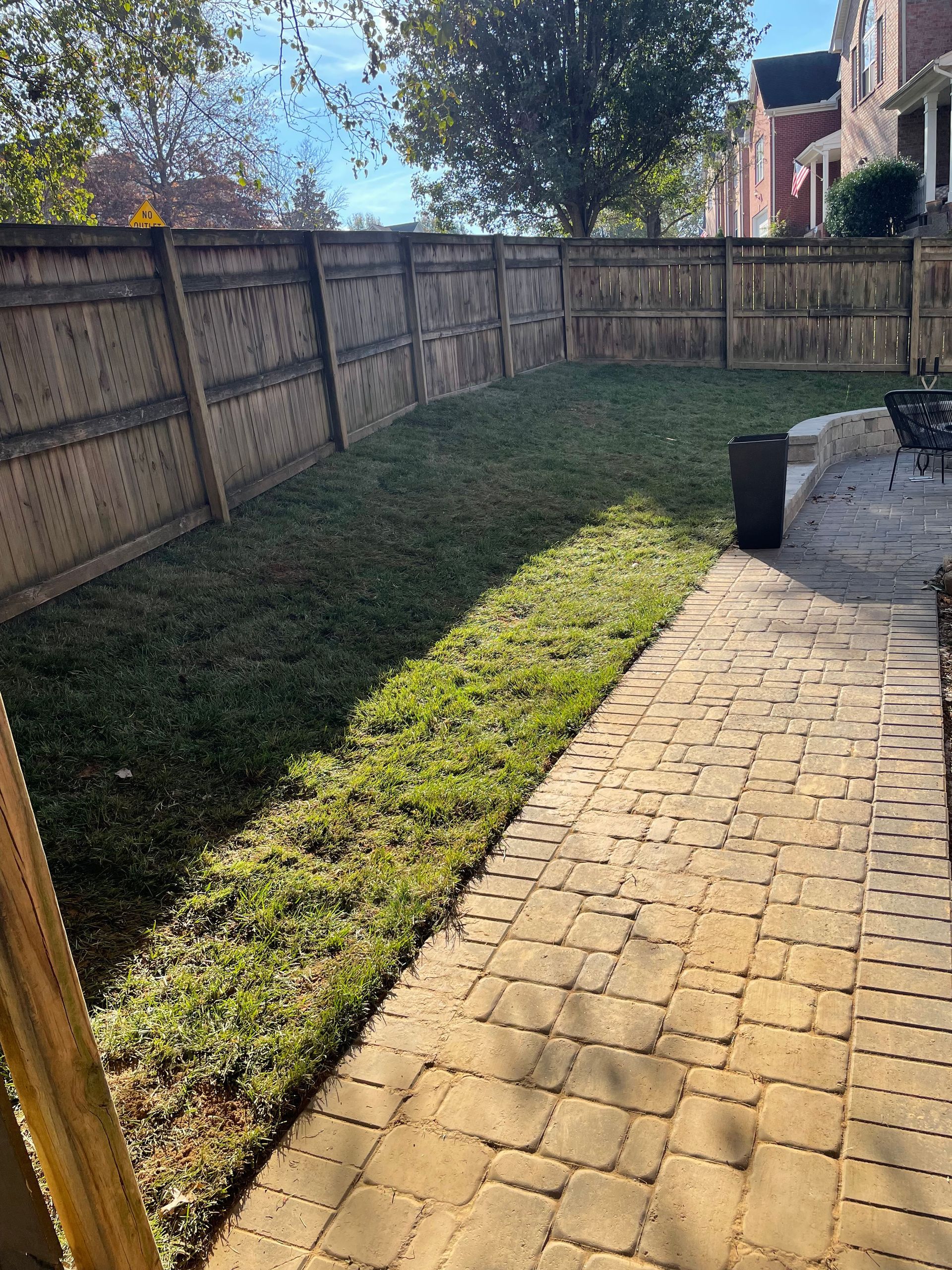Backyard with wooden fence, brick patio, and grassy area; sunny day.