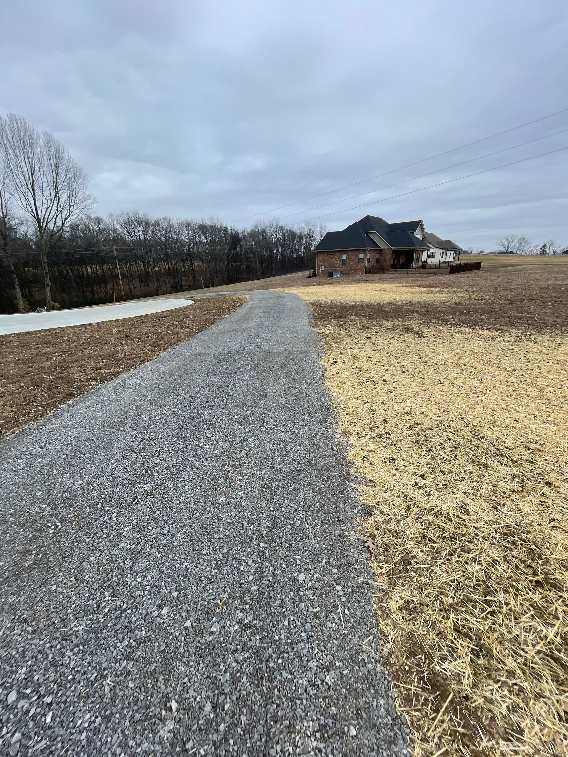 Gravel path leads toward a house on a cloudy day. Light brown and yellow fields flank the path.
