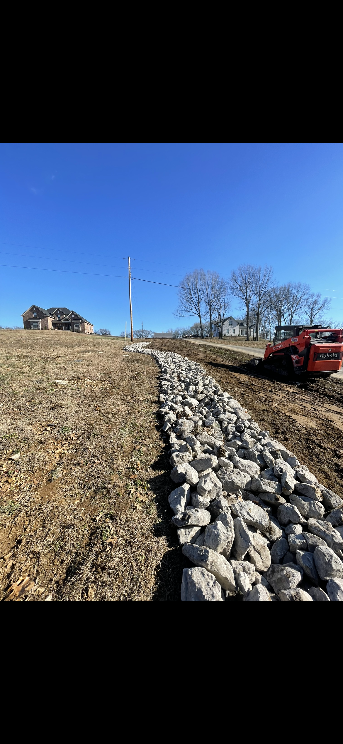A rock-lined ditch with a small excavator, next to a house under construction. Bright blue sky.