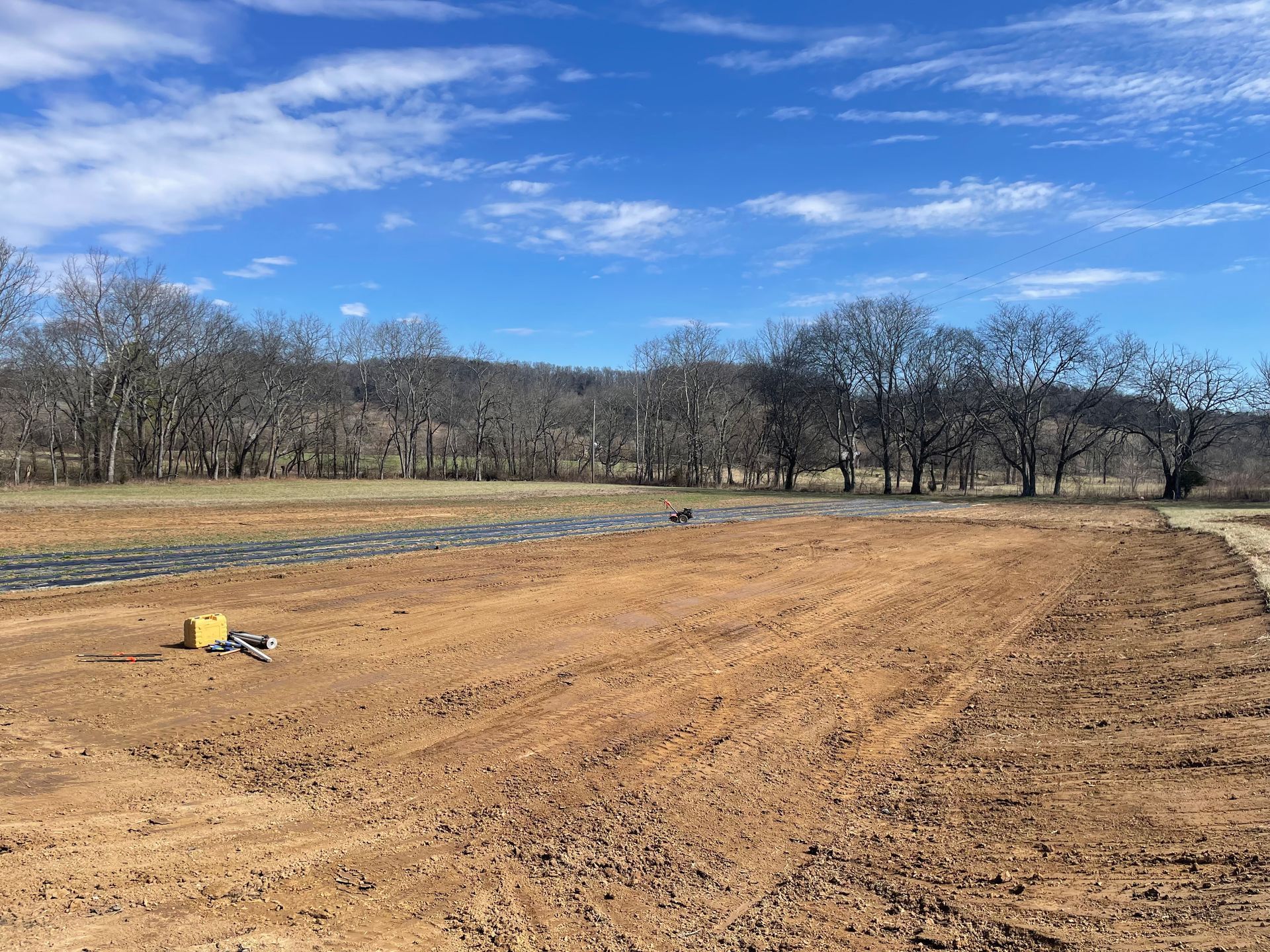 Brown field under a blue sky. A small machine and a covered area are on the field's surface with trees in the background.