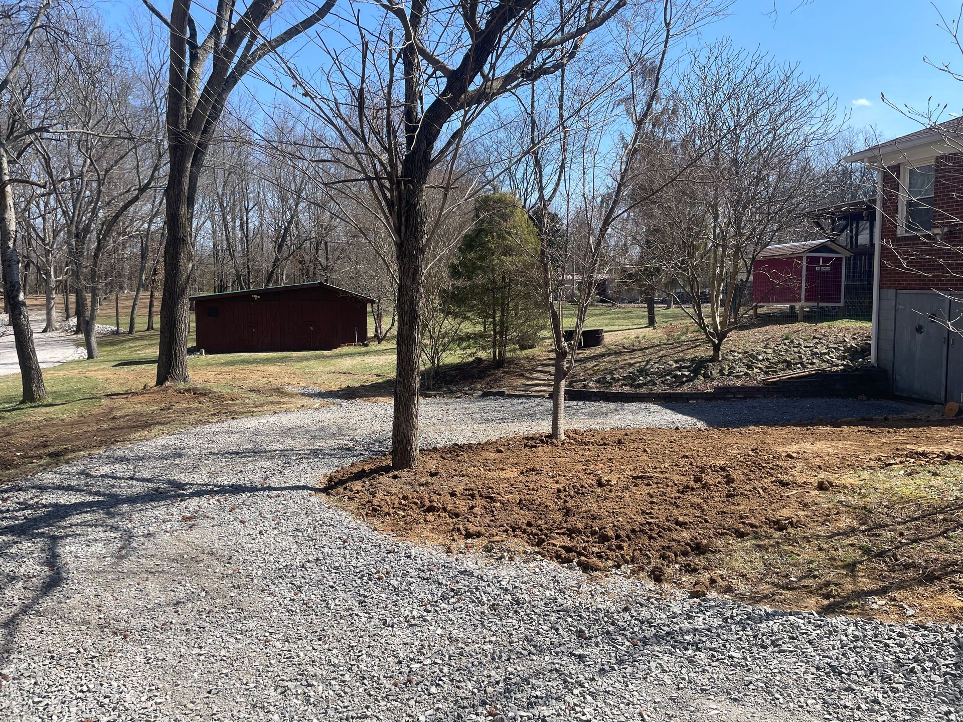 Gravel driveway with a shed and red building on a sunny day with trees.