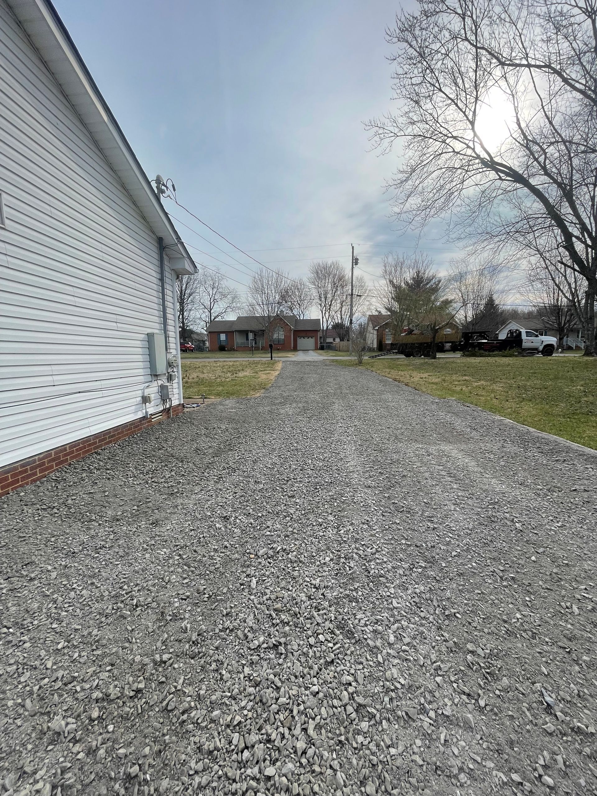 Gravel driveway leading to a brick building under a cloudy sky, beside a white house with a tree.