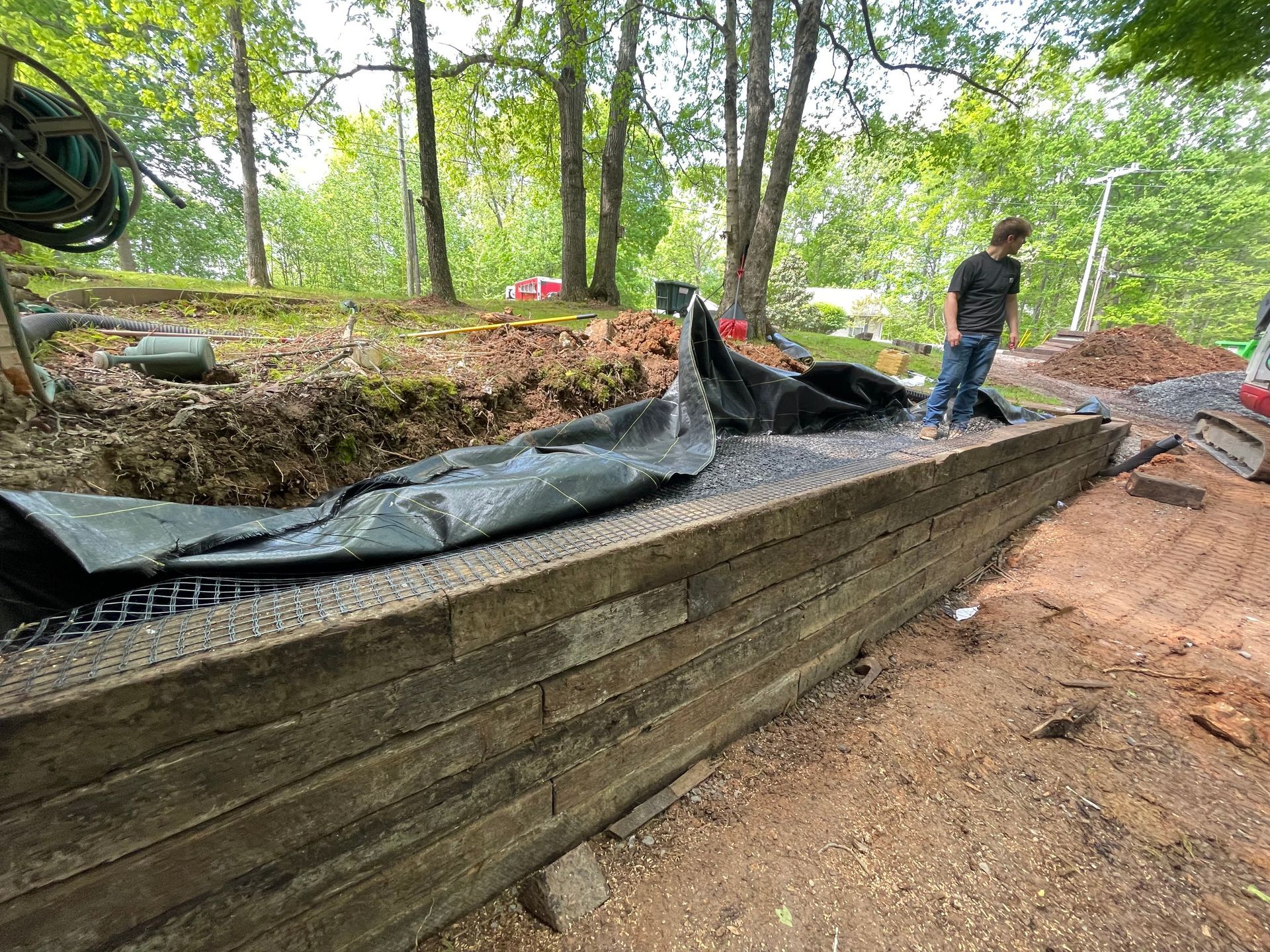 Retaining wall under construction. Person standing near the wall with black mesh and soil. Forest in the background.