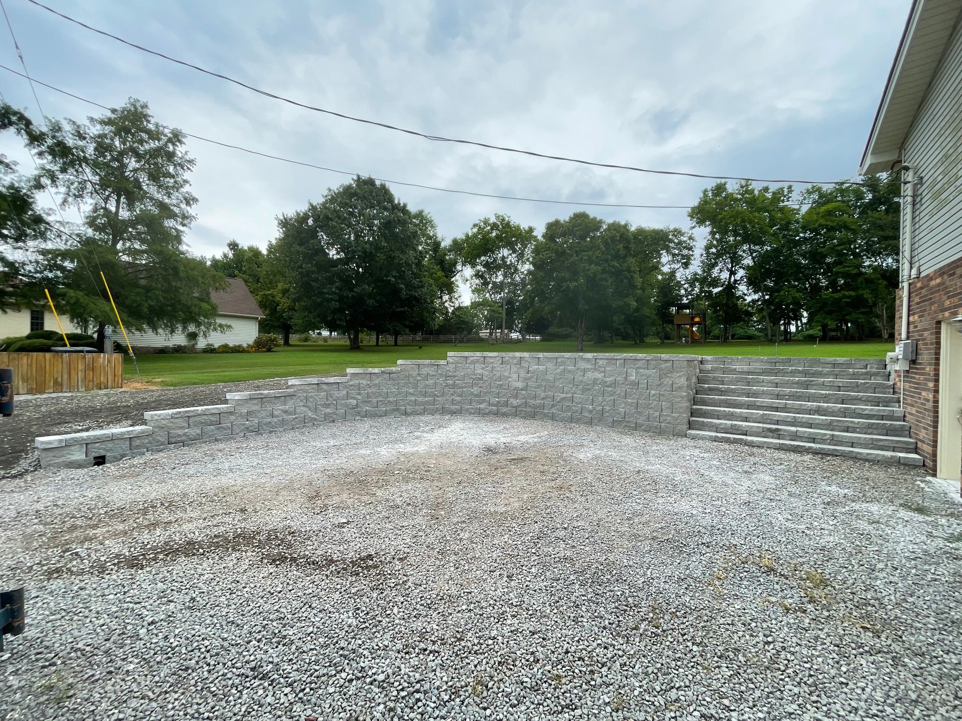 Gravel driveway with retaining wall, steps, and a house on the right, leading up to a grassy area under a cloudy sky.